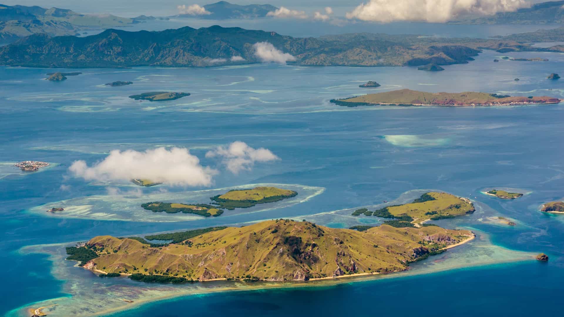 A stunning high-angle aerial view of Komodo Island and Batu Moncho in Indonesia, featuring lush green islands surrounded by vibrant turquoise waters and visible coral reefs under a bright sky.