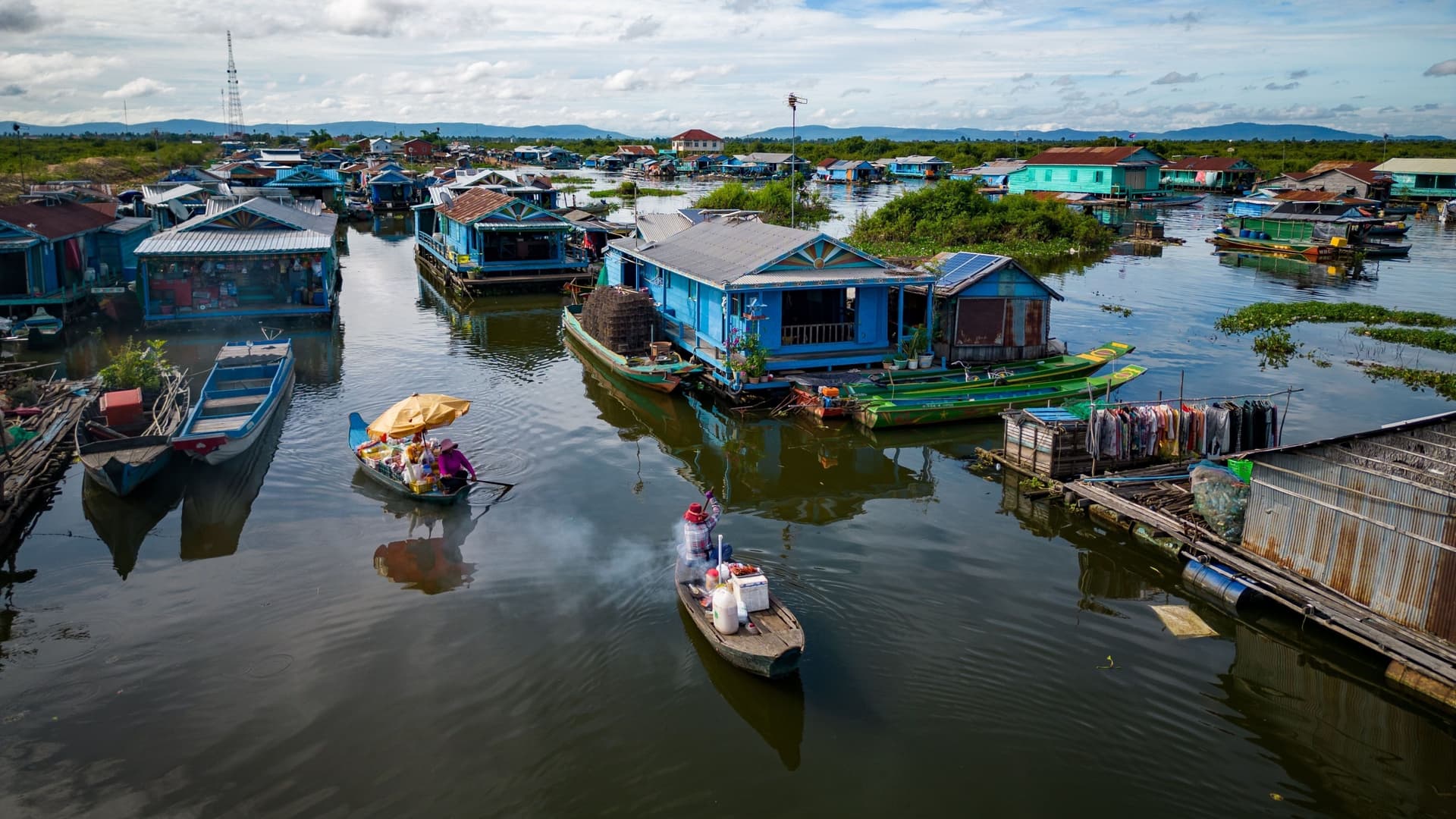 A wide-angle view of the bustling floating village of Kompong Luong in Cambodia, with colorful houses and boats on the water and mountains in the background.