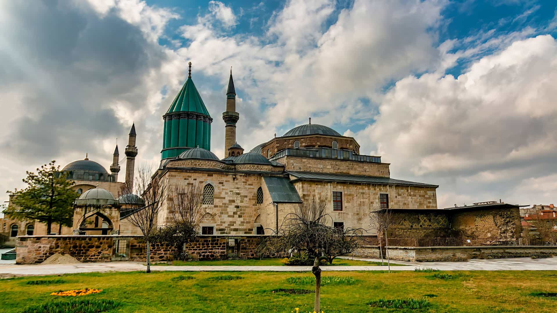 "A panoramic shot of the historic Mevlana Museum in Konya, Turkey, with its famous fluted green dome and multiple minarets under a dramatic cloudy sky, with a green grassy lawn in the foreground. "