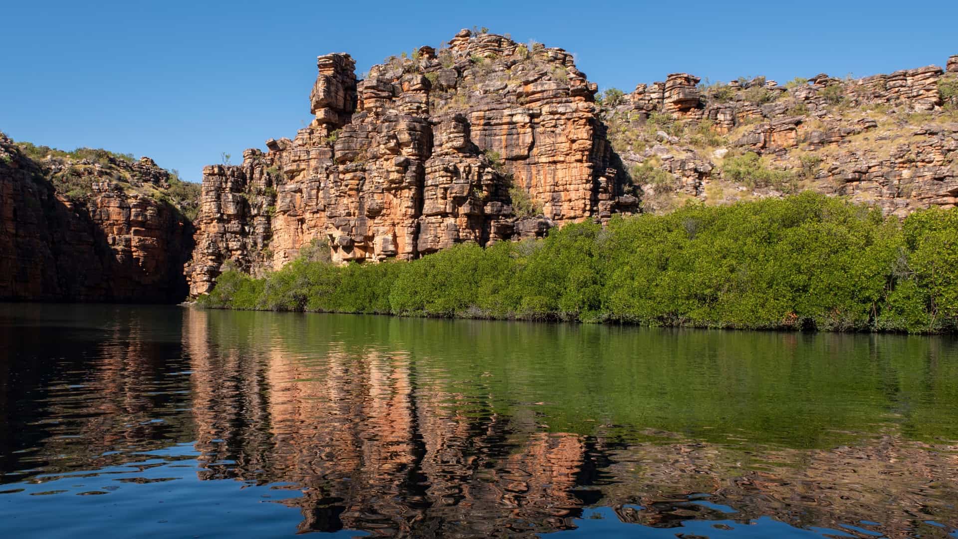 A tranquil river scene at Koolama Bay, Australia, with stunning red and orange-layered cliffs and lush green mangroves reflecting perfectly in the calm water under a clear blue sky.