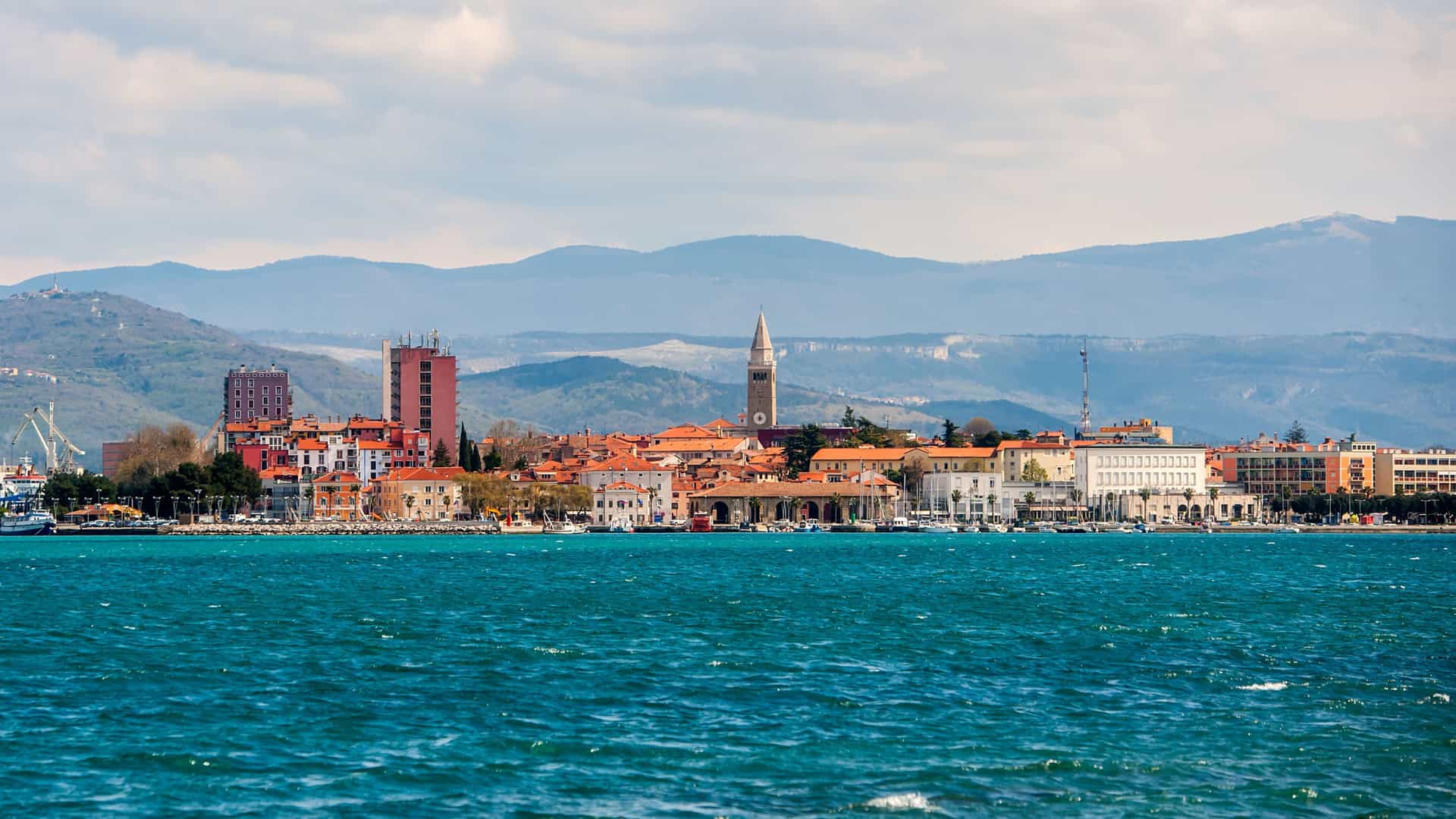 A vibrant panoramic view of Koper, Slovenia, with a beautiful waterfront, historic red-roofed buildings, and a church steeple, set against a backdrop of rolling mountains.