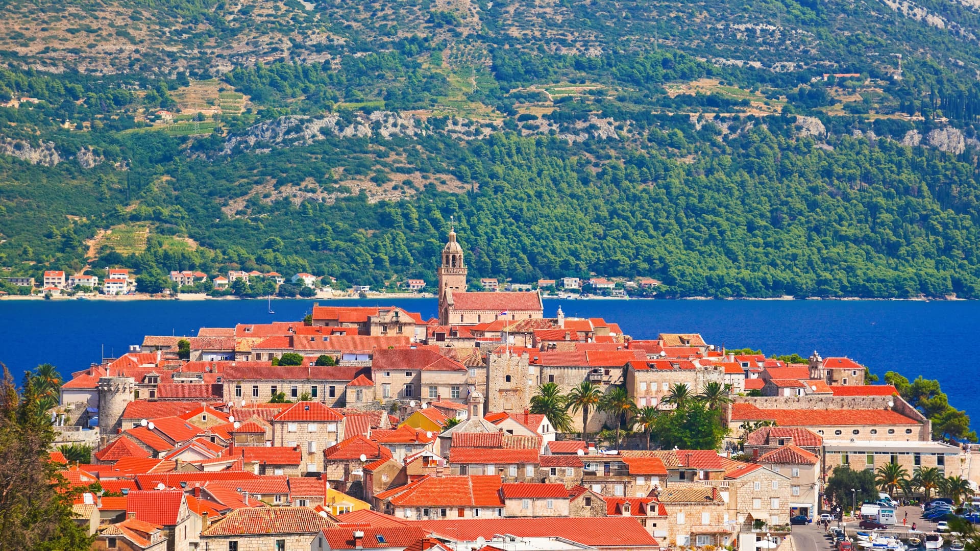 A scenic view of Korcula Old Town in Croatia, featuring red-tiled roofs, a church steeple, and a historic stone wall set against the deep blue Adriatic Sea and lush green mountains.