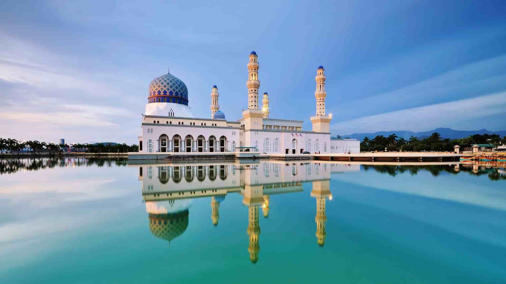 A wide shot of the beautiful Kota Kinabalu City Mosque in Malaysia, featuring its majestic white and blue structure reflecting perfectly on the serene, glassy water surrounding it at sunset.