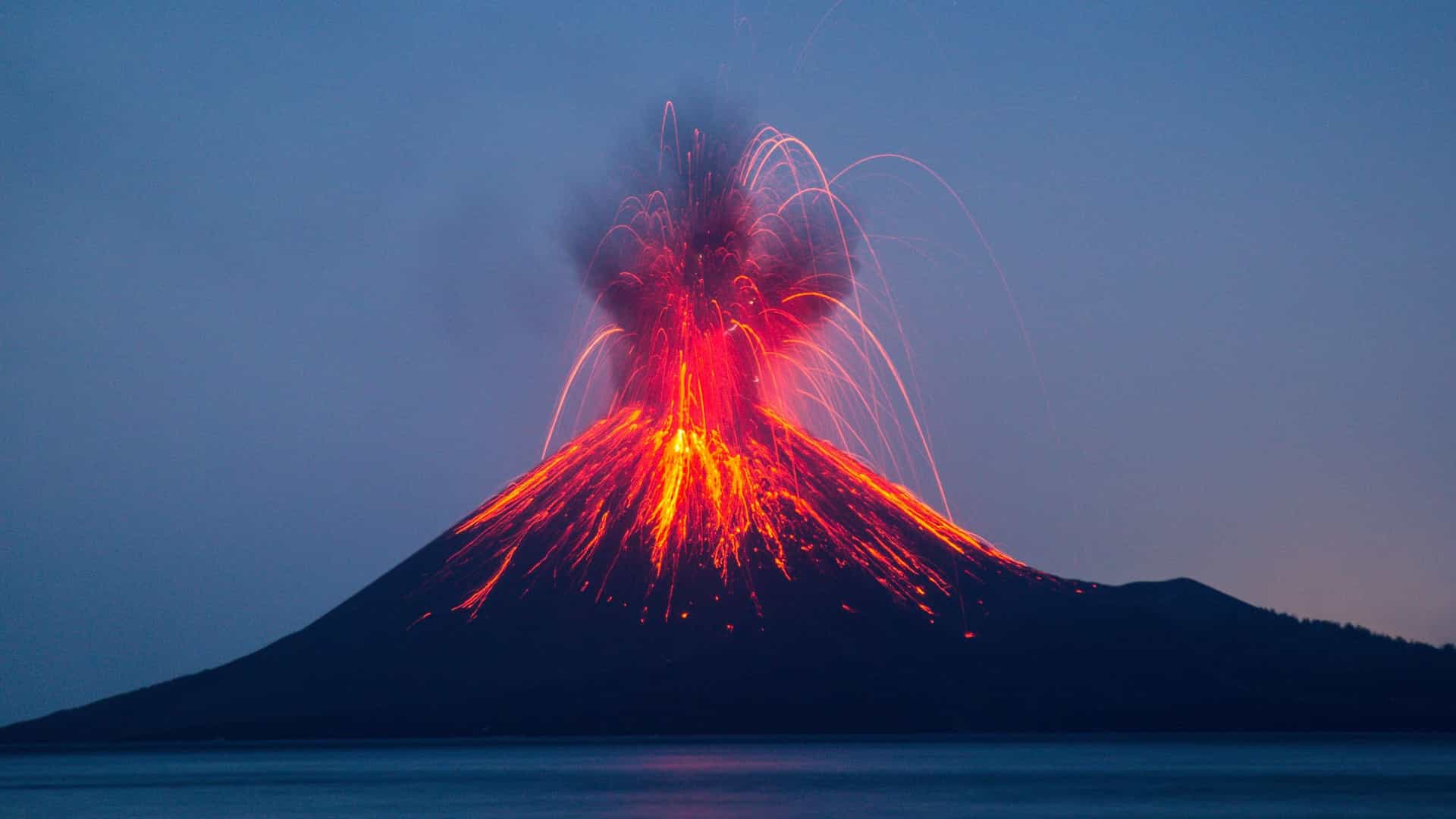 A nighttime long exposure shot of the Anak Krakatau volcano in Indonesia erupting, with bright red lava and sparks streaming down the mountain and shooting into the dark blue sky.