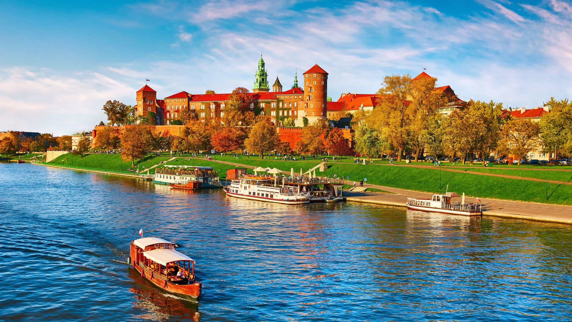 "A vibrant autumn view of Wawel Royal Castle in Krakow, Poland, with its red brick walls and towers, situated on a hill overlooking the Vistula River with several tour boats. "