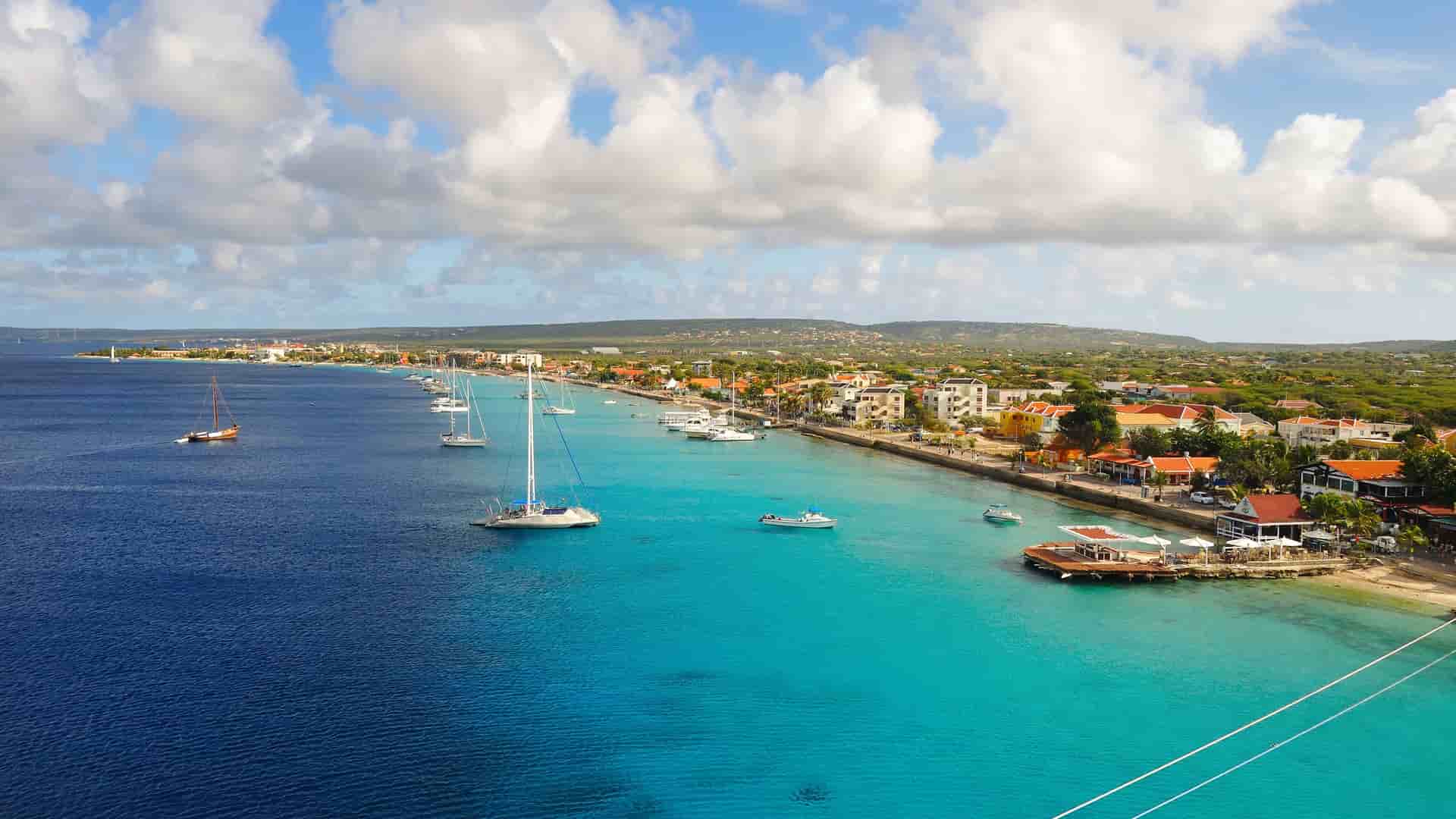 An aerial view of the Kralendijk waterfront in Bonaire, showcasing the contrast between the deep blue ocean and the shallow turquoise water near the shore, with sailboats anchored offshore.