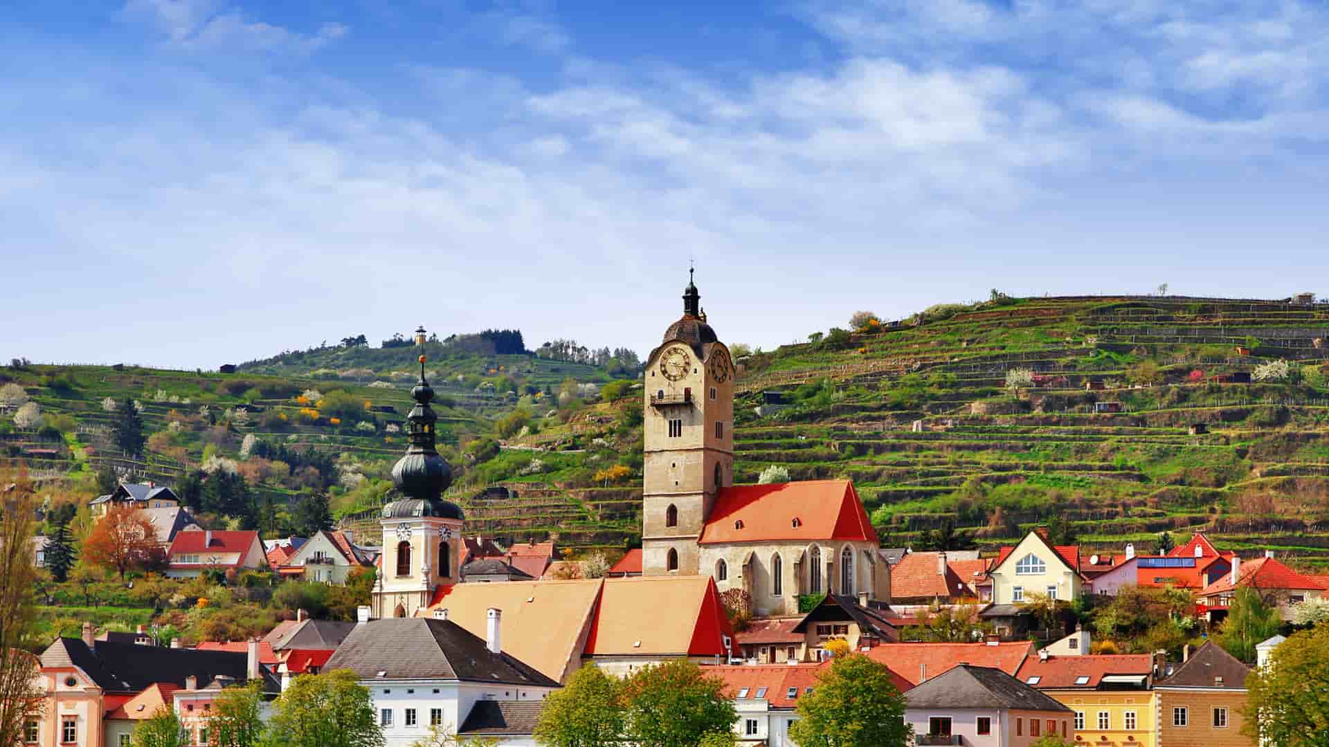 A scenic view of Krems an der Donau in Austria, showcasing the historic old town buildings with a prominent church tower, set against lush green, terraced hillsides and a blue sky.
