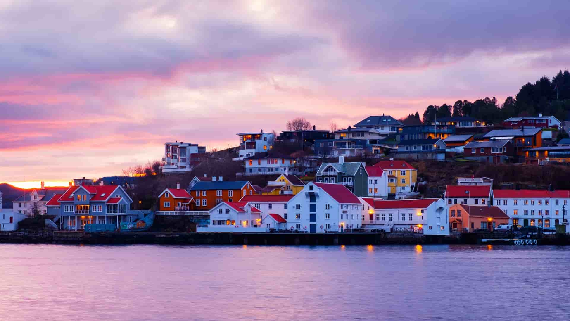 A colorful array of houses on the waterfront of Kristiansund, Norway, with the warm lights of the buildings reflecting in the water during a beautiful pink and purple sunset.