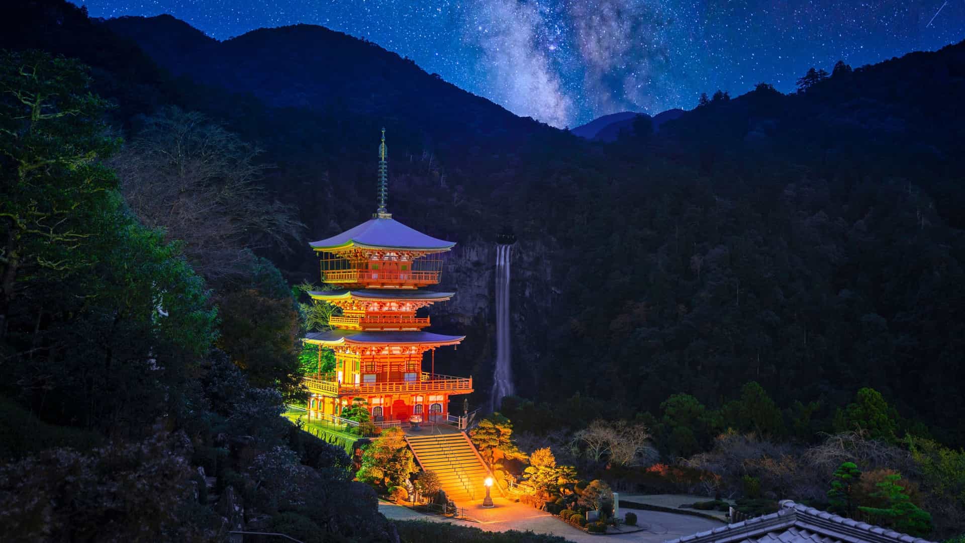 A nighttime view of the vibrant, illuminated Nachi Falls and the iconic three-story pagoda of Seiganto-ji temple in Kumano, Japan, with the Milky Way visible in the night sky.