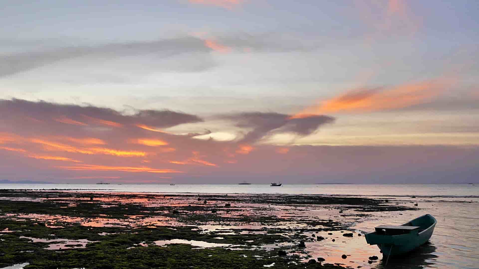 A breathtaking sunset over a tidal flat in Kupang, West Timor, Indonesia, with a small fishing boat on the shore and the sky filled with vibrant orange and purple clouds.