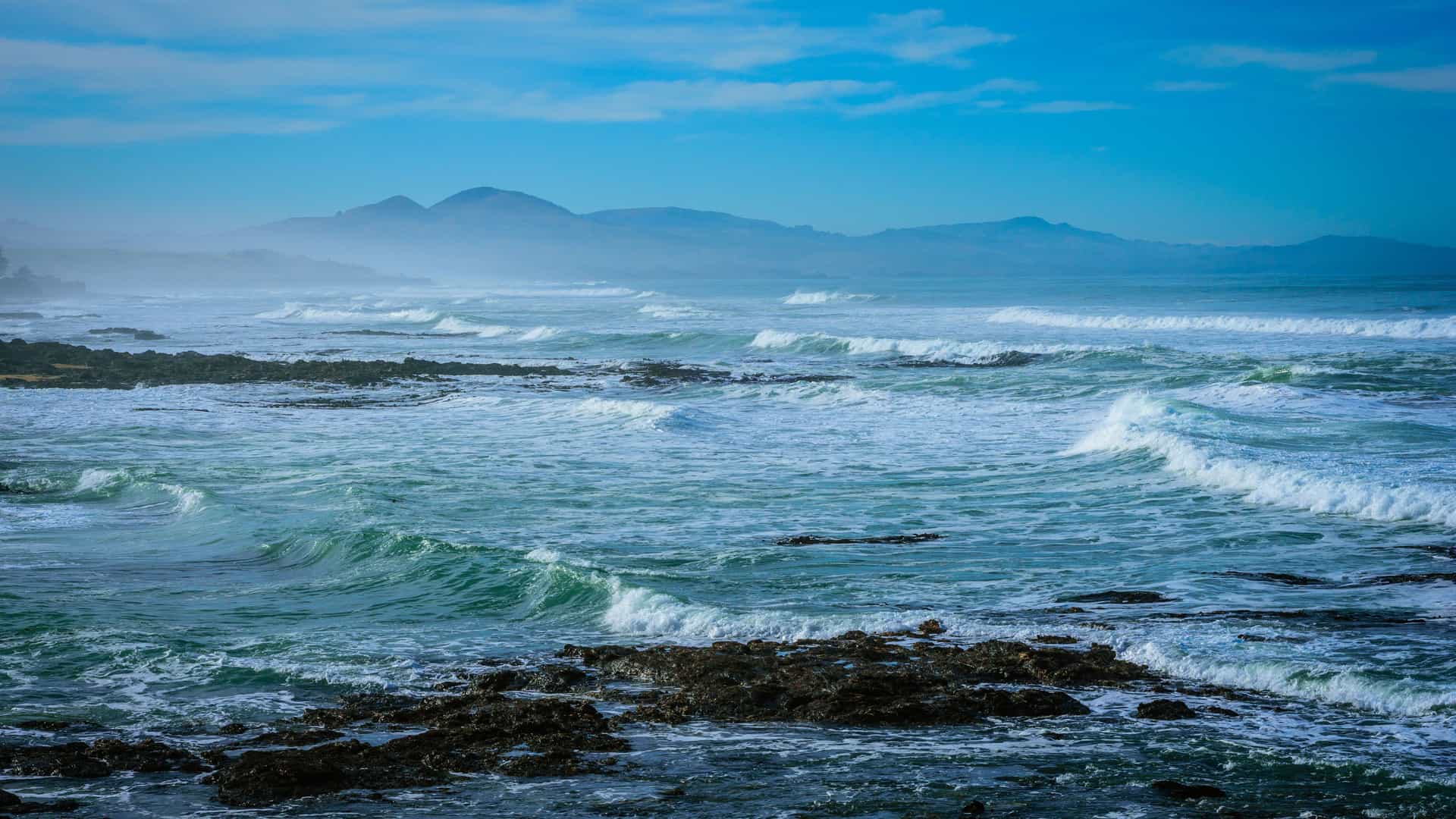 A stunning view of the rugged coastline of Kuri Bay in Western Australia, with foamy turquoise waves crashing on the rocky shore and misty hills visible in the distance under a blue sky.