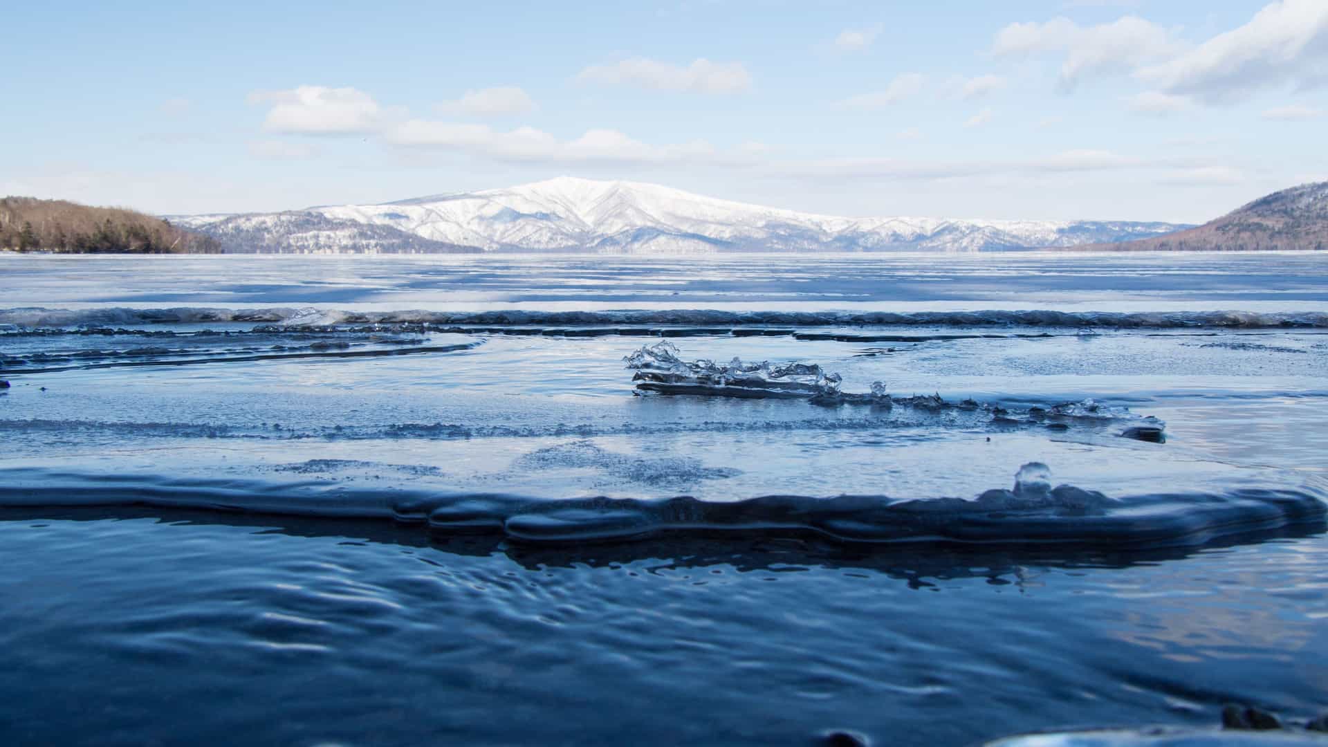 A scenic winter view of Lake Akan in Kushiro, Japan, with a layer of thin ice on the water in the foreground and snow-covered mountains visible in the distance under a blue sky.