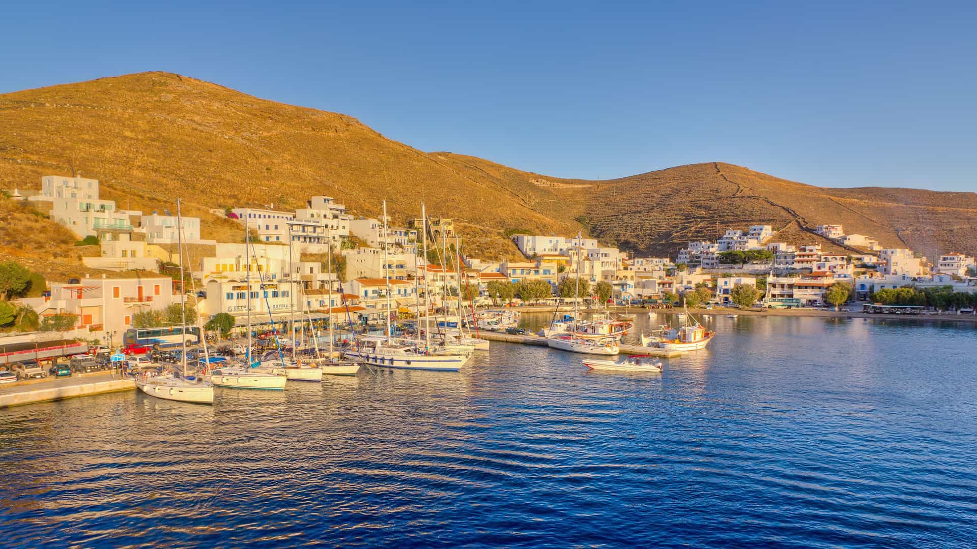 "A picturesque panoramic view of the Kythnos port in Greece, with white Cycladic houses and buildings lining the waterfront and a marina filled with sailboats, all nestled against the backdrop of dry, golden hills under a clear blue sky. "