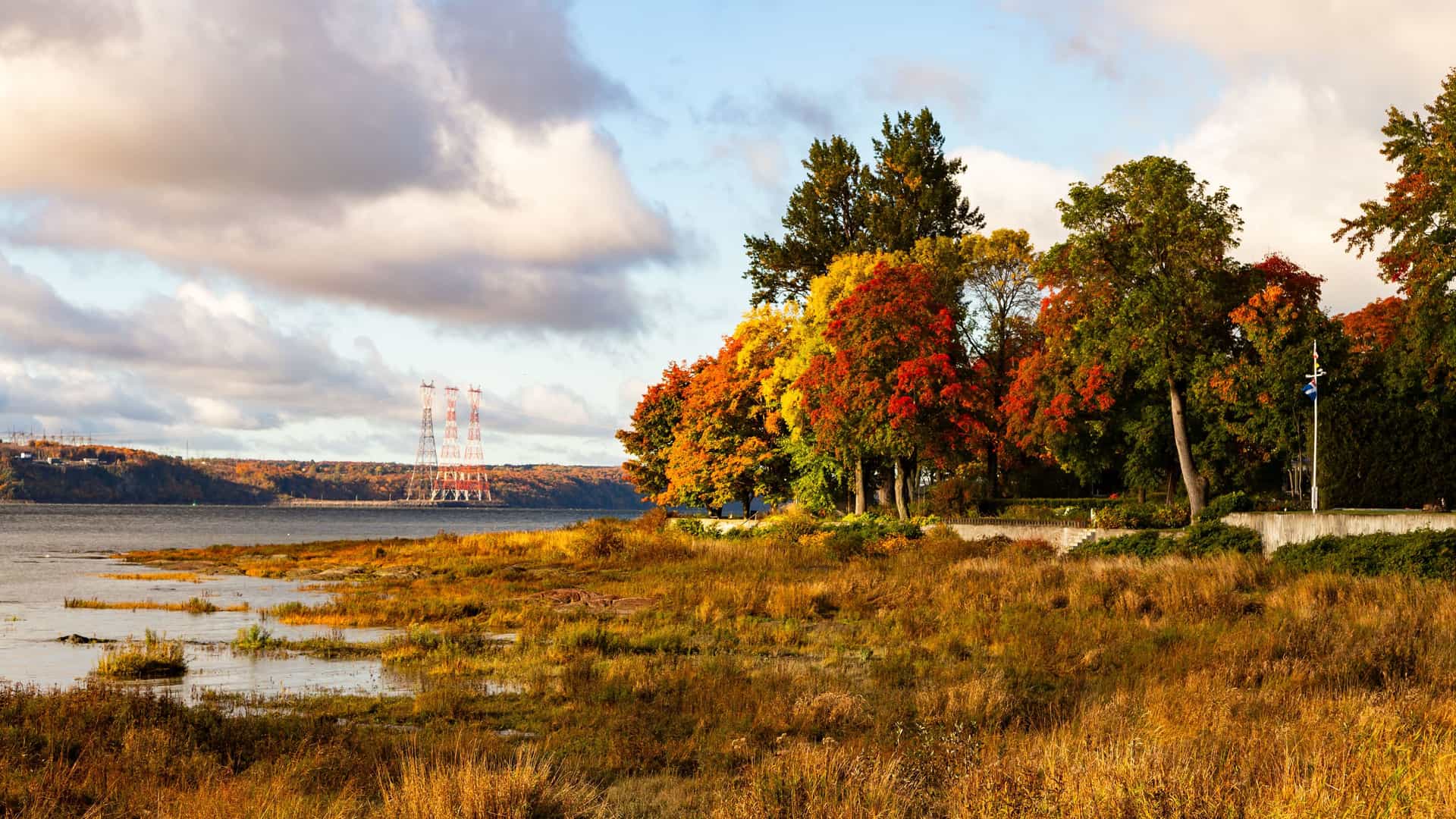 "A wide-angle landscape shot of the St. Lawrence River in L'Anse-au-Clair, Canada, showing a shoreline of marshy grass and a line of trees with vibrant autumn foliage. "
