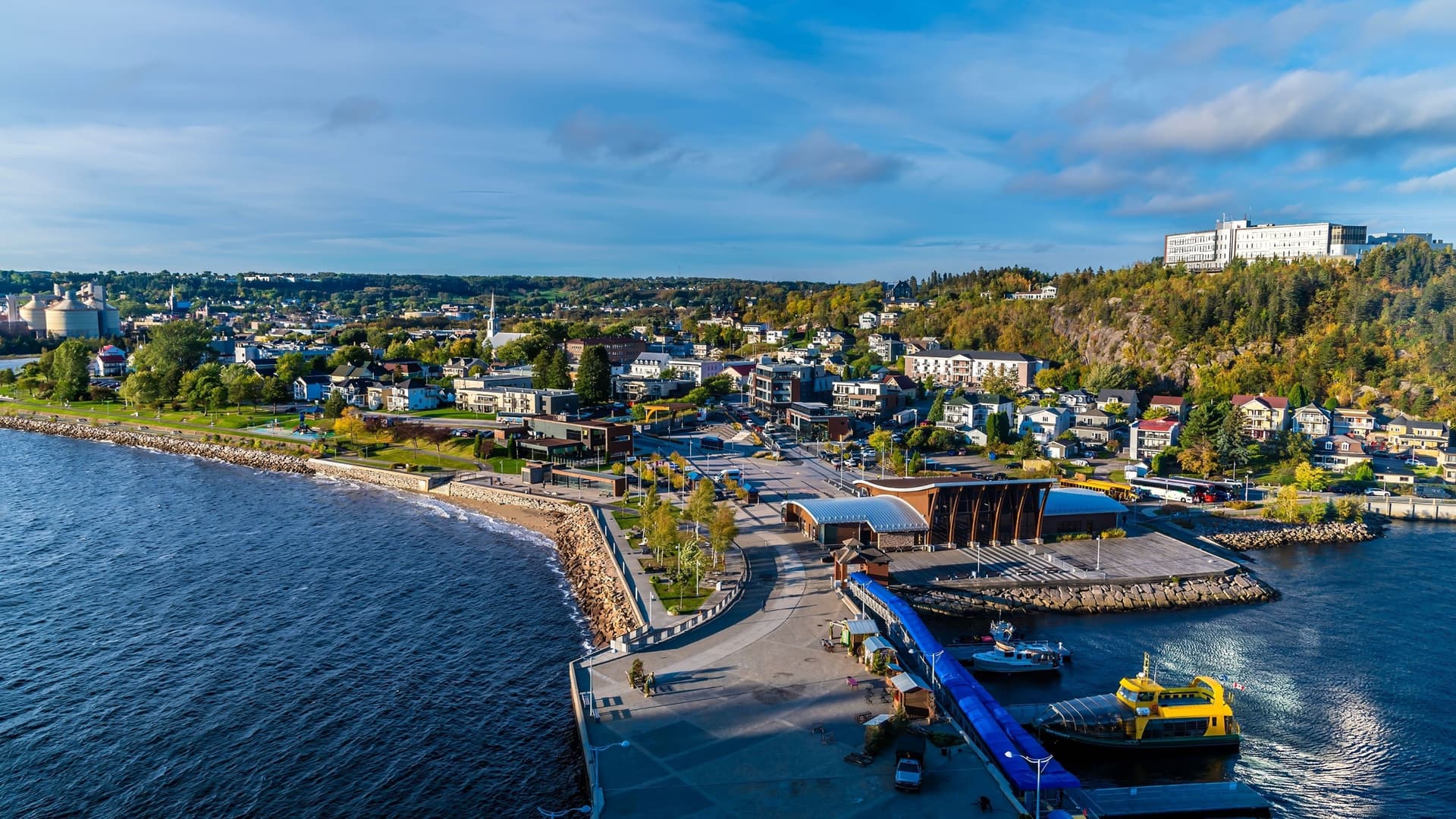 An aerial view of the scenic La Baie waterfront in Saguenay, Quebec, with a marina, city buildings, and residential homes against a backdrop of fall foliage on the hillside.