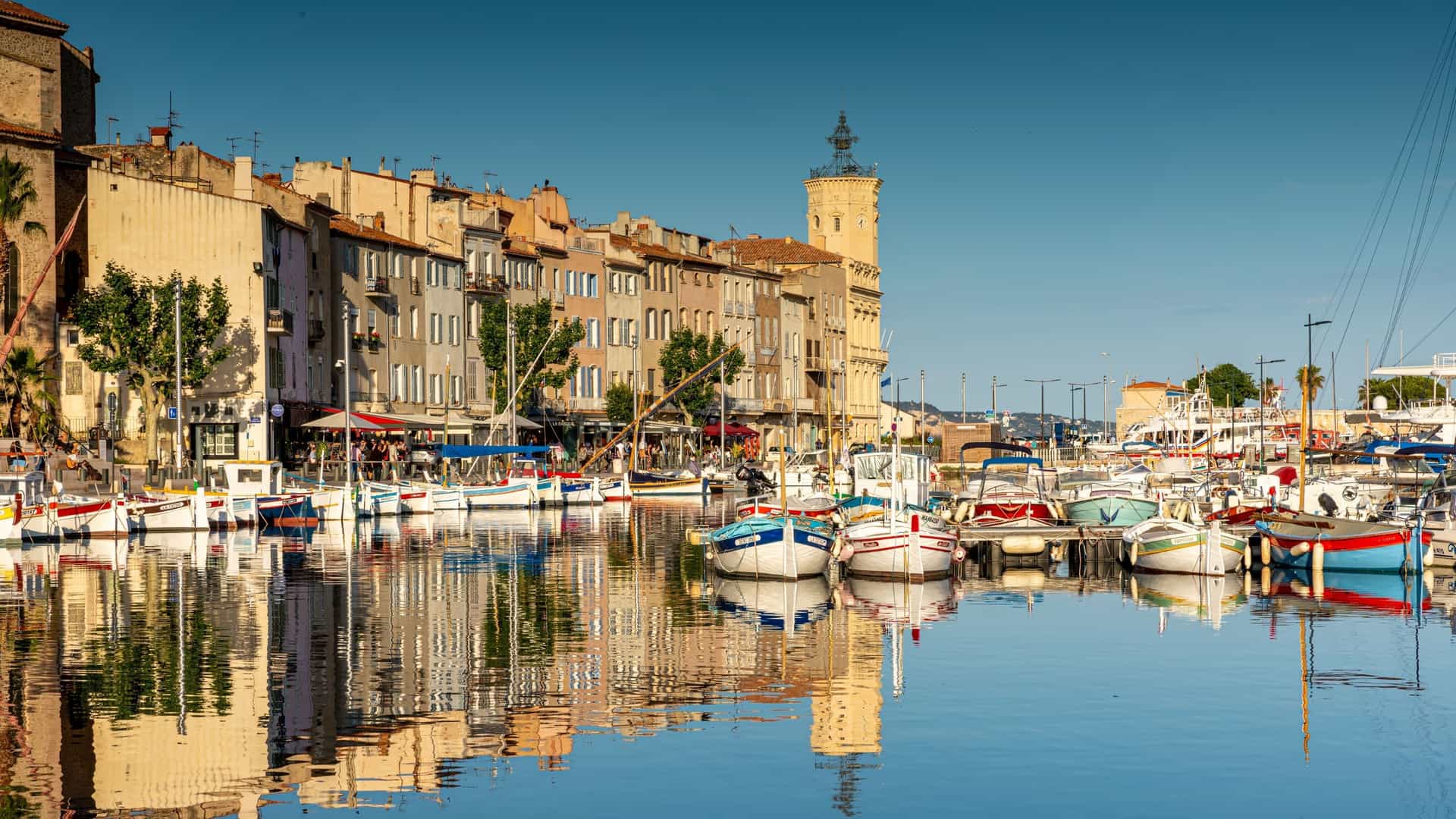 A scenic view of the charming harbor of La Ciotat, France, with colorful traditional fishing boats docked in the calm water reflecting the historic buildings and a prominent clock tower.