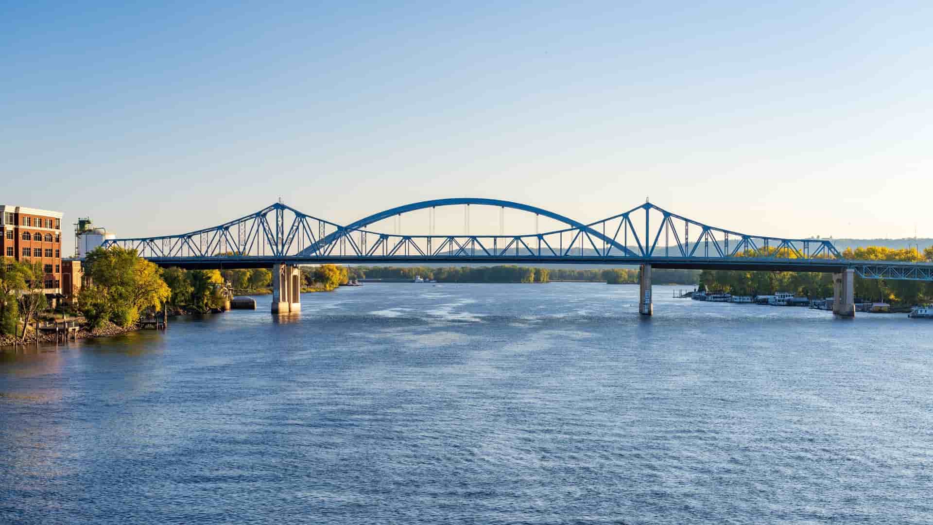 A long-shot view of the La Crosse High Bridge spanning the Mississippi River on a clear day, with trees lining the banks and a building visible on the far left.