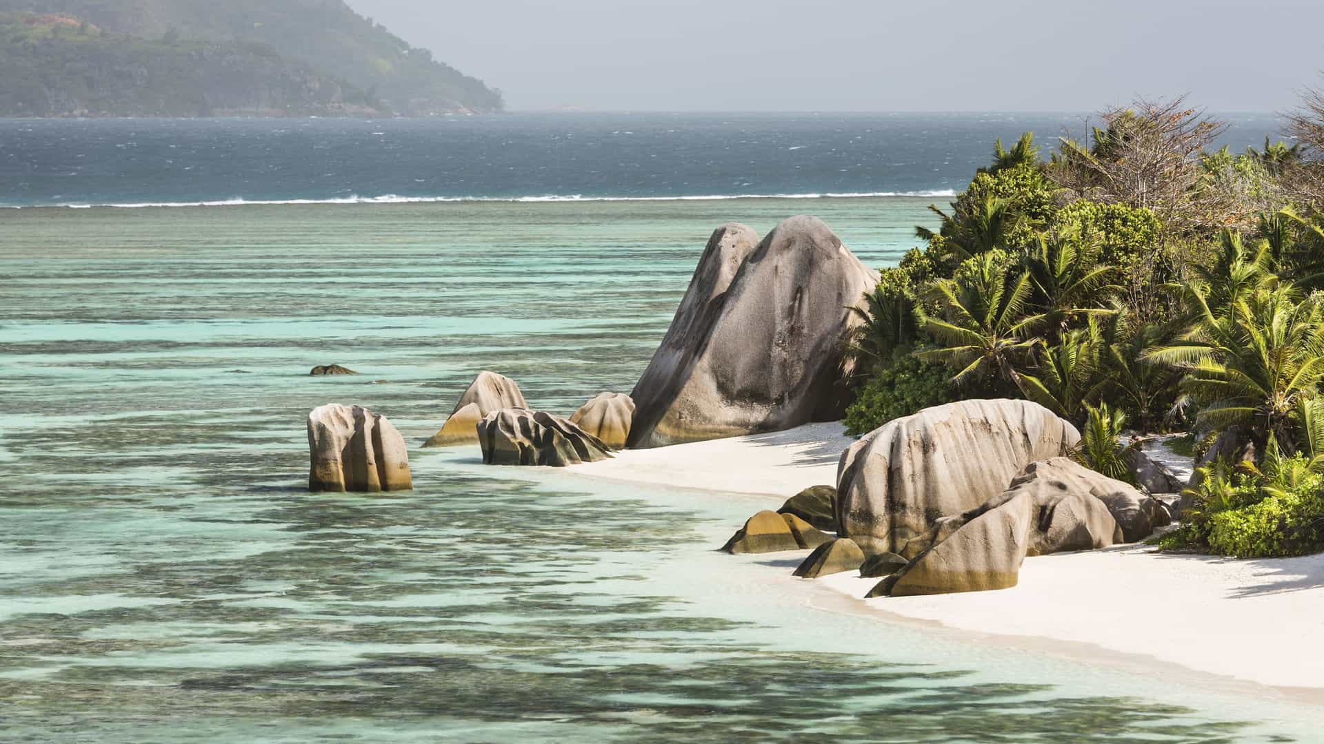 A scenic wide-shot of the Anse Source d'Argent beach in La Digue, Seychelles, showcasing its stunning white sands, iconic granite boulders, and clear turquoise waters.