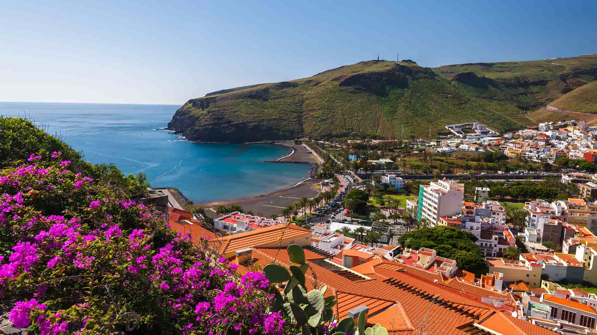 A high-angle view of the coastal town of San Sebastián de La Gomera, with red-tiled roofs, a black-sand beach, and a deep blue bay framed by bougainvillea flowers and verdant hills under a clear sky.