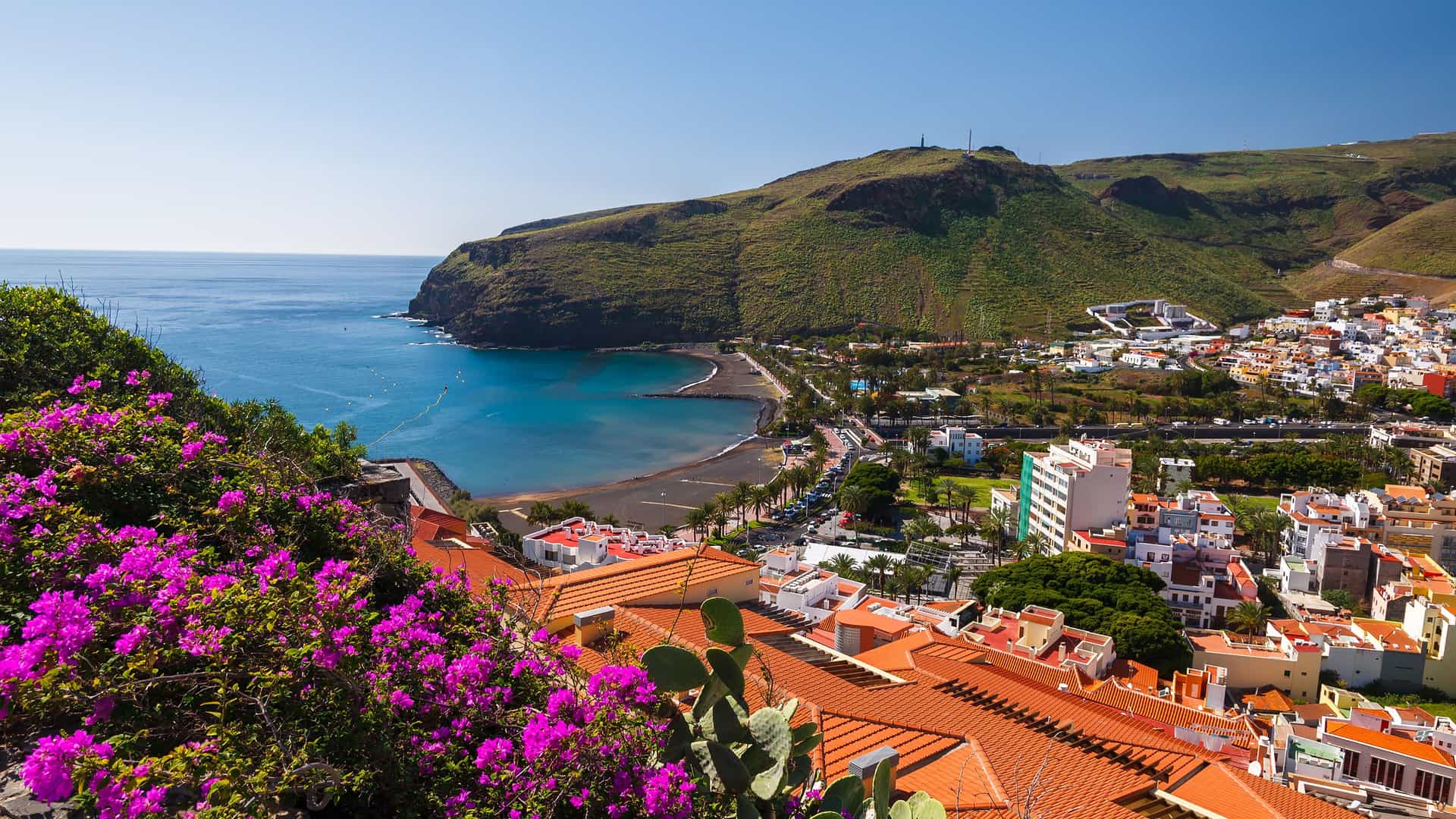 A high-angle view of the coastal town of San Sebastián de La Gomera, with red-tiled roofs, a black-sand beach, and a deep blue bay framed by bougainvillea flowers and verdant hills under a clear sky.