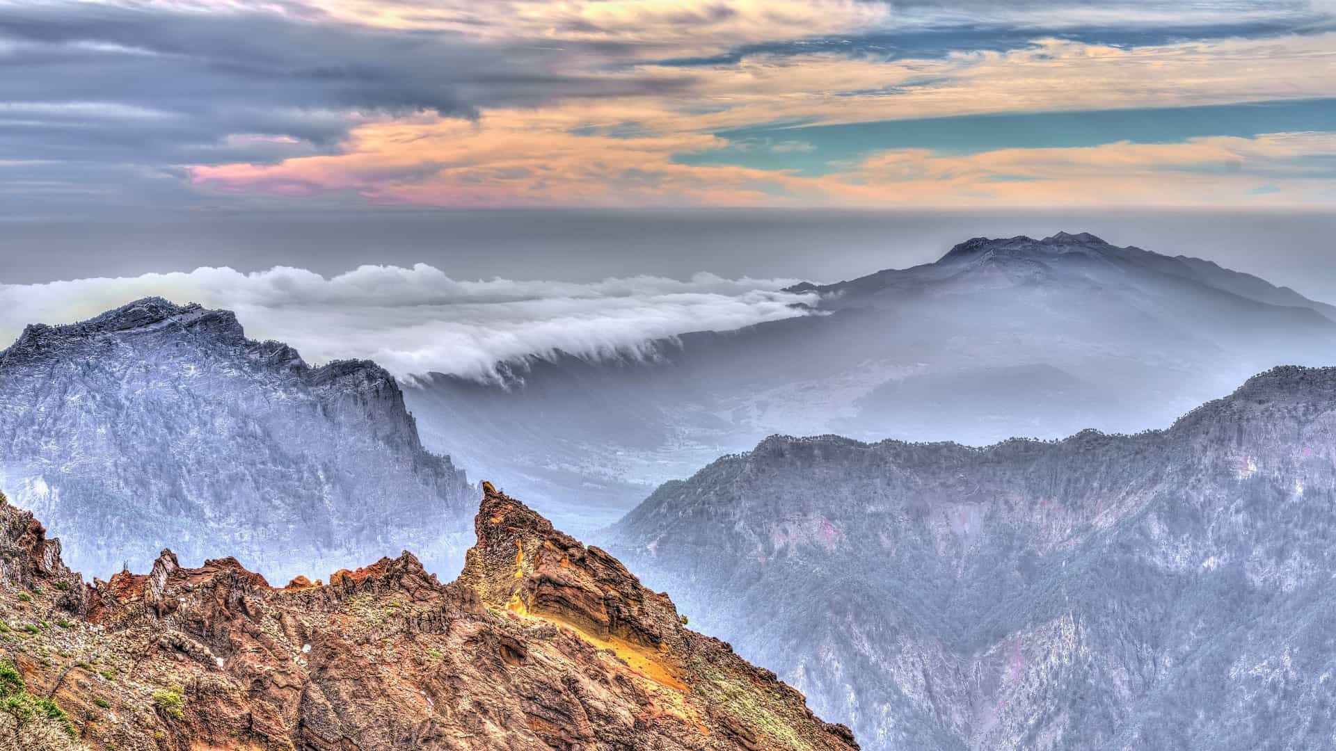 A high-angle view of the dramatic, rugged volcanic mountains of La Palma, with clouds flowing through the valleys and the ocean visible on the horizon under a colorful sky.