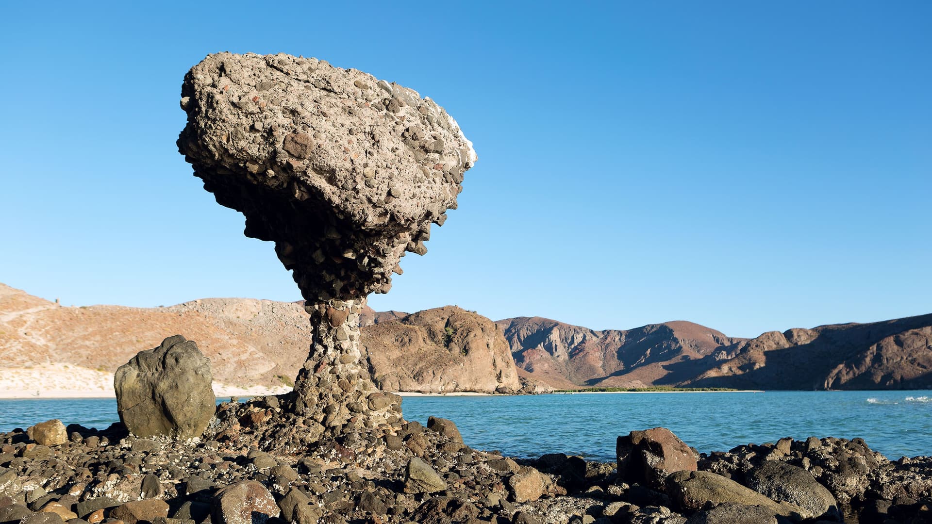 A striking mushroom-shaped rock formation, known as a "hongos," stands on the rocky beach of Balandra Bay near La Paz, Mexico, with calm turquoise water and arid mountains in the background.
