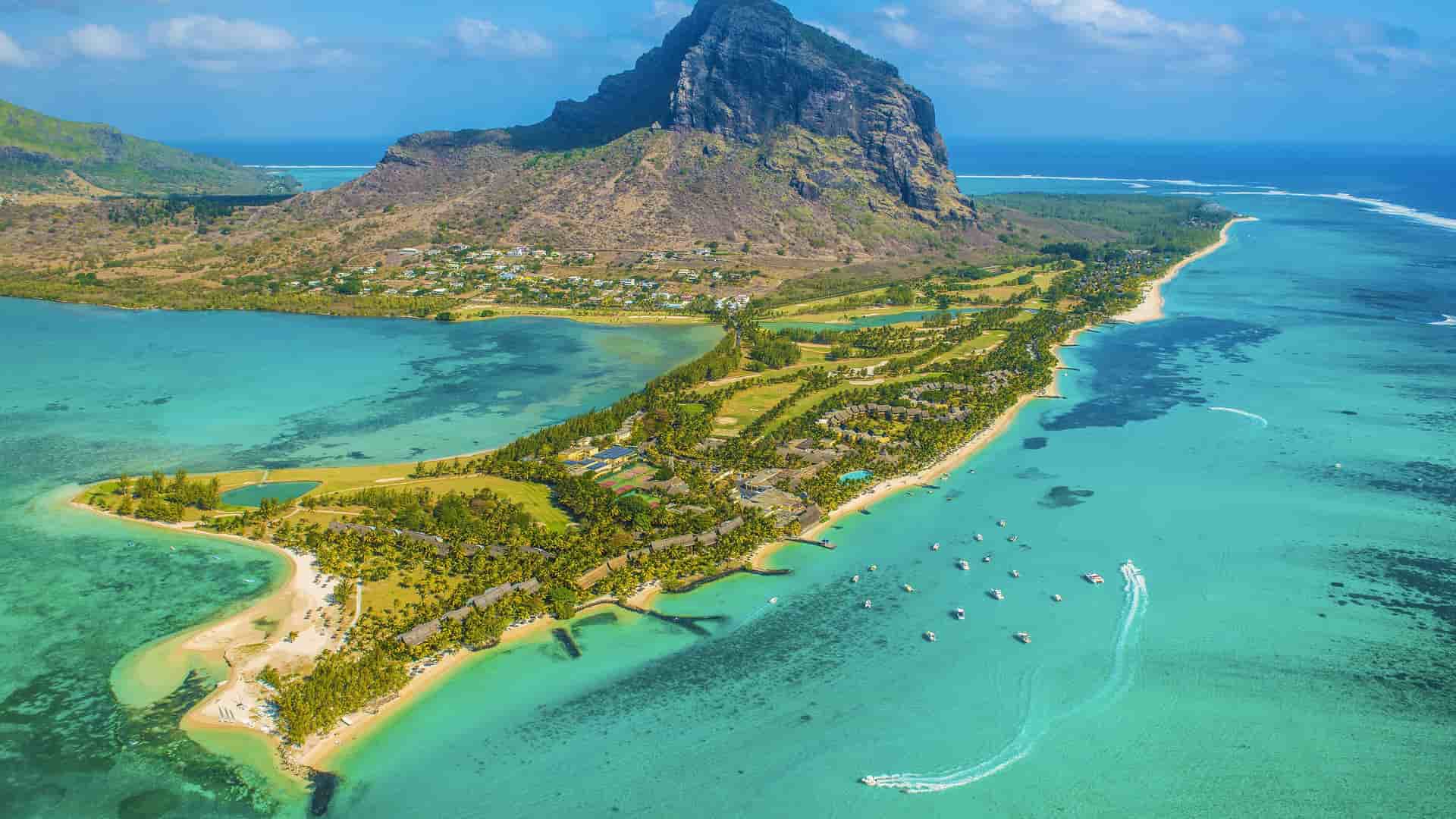 An aerial view of a luxurious resort and golf course on a peninsula in La Possession, Réunion, with a majestic volcanic mountain rising in the background and a turquoise lagoon surrounding the coastline.
