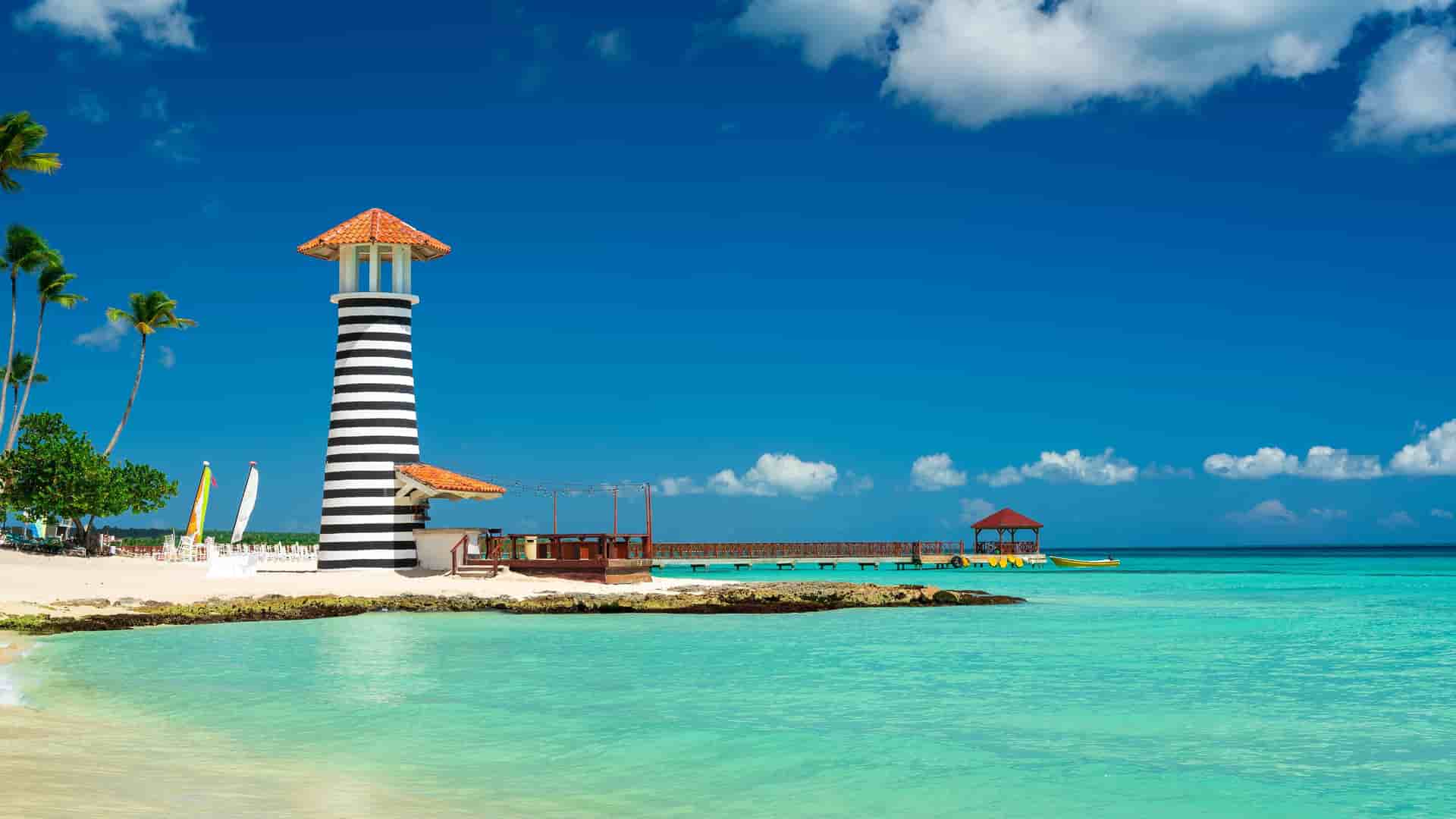 "A picturesque white sand beach in La Romana, Dominican Republic, with a distinctive black and white striped lighthouse and a wooden pier extending over the clear turquoise water. "
