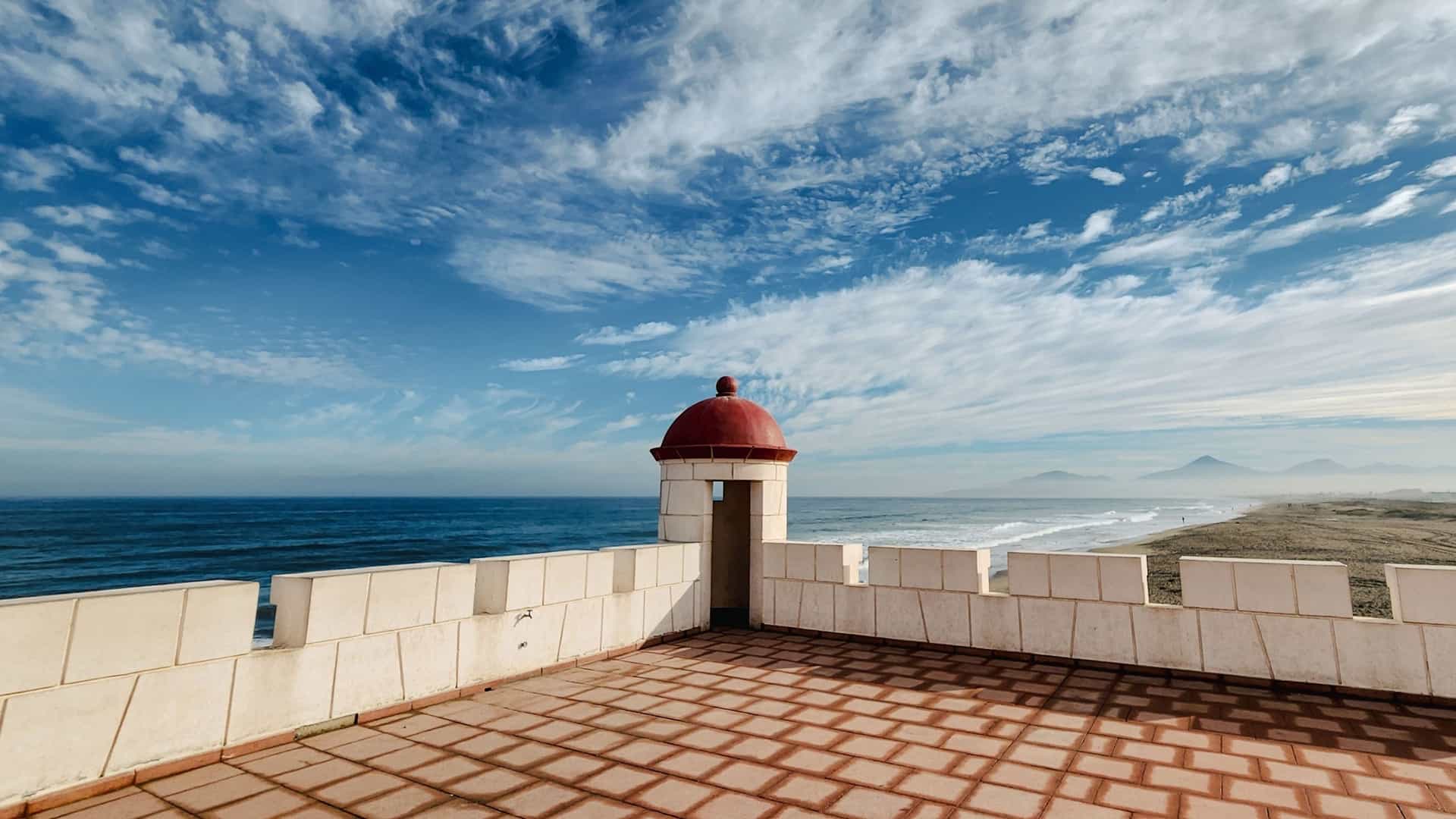 A sunlit terrace with a red-domed turret overlooking the Pacific Ocean and a wide sandy beach in La Serena, Chile.