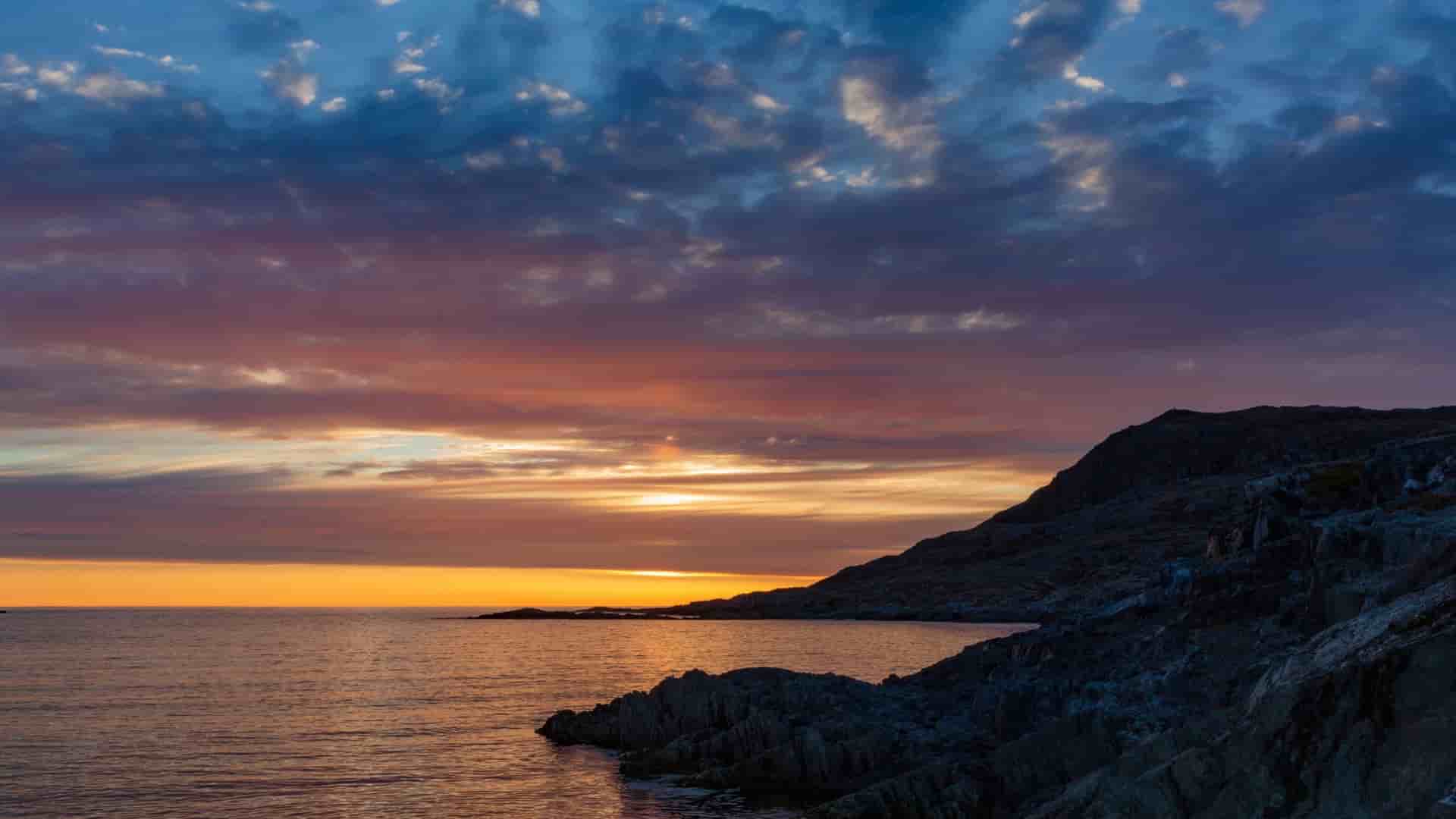 A beautiful sunset over the Labrador Sea, with a vibrant orange and blue sky reflected on the water and a rugged rocky coastline in silhouette.