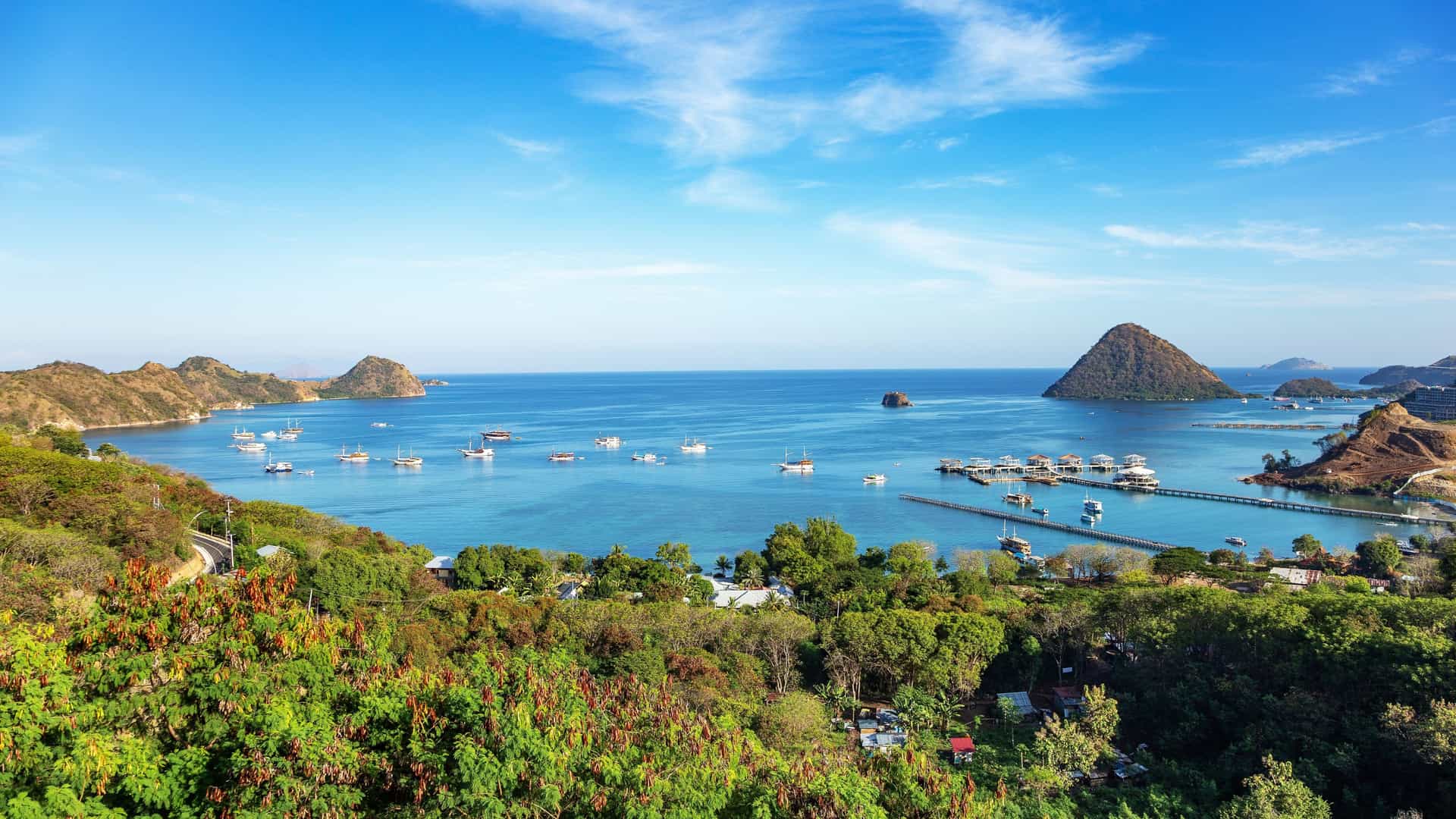 A wide, scenic view of Labuan Bajo in Indonesia, featuring clear blue water with several traditional boats anchored in the harbor, surrounded by lush green mountains under a bright, sunny sky.