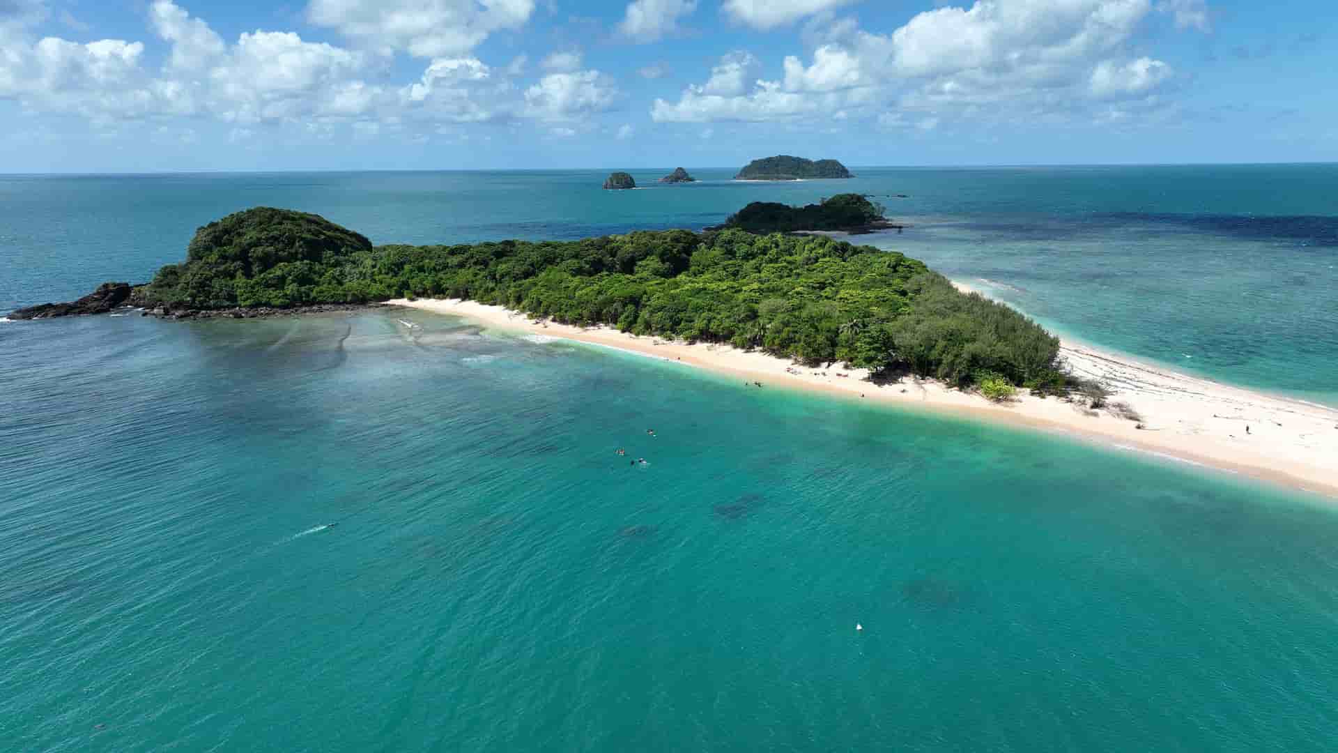 An aerial view of Lady Franklin Island, a remote tropical island with a sandy beach and lush green forest, surrounded by turquoise water under a blue sky with clouds.
