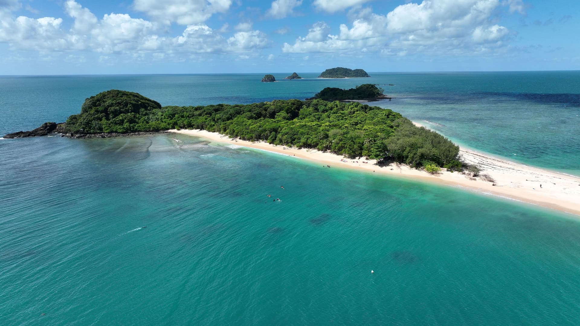 An aerial view of Lady Franklin Island, a remote tropical island with a sandy beach and lush green forest, surrounded by turquoise water under a blue sky with clouds.