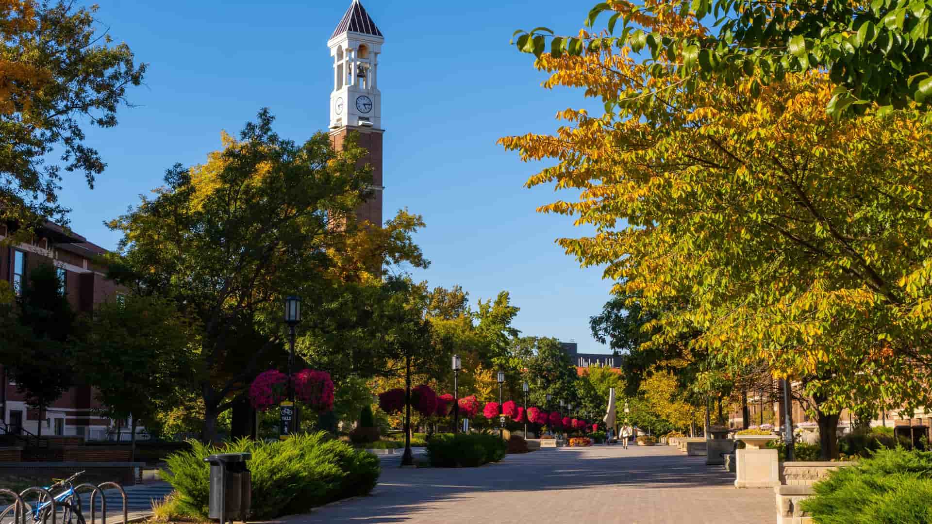 "A vibrant autumn scene on the campus of Purdue University in Lafayette, Indiana, showing the iconic Bell Tower and a path lined with trees in full fall foliage. "