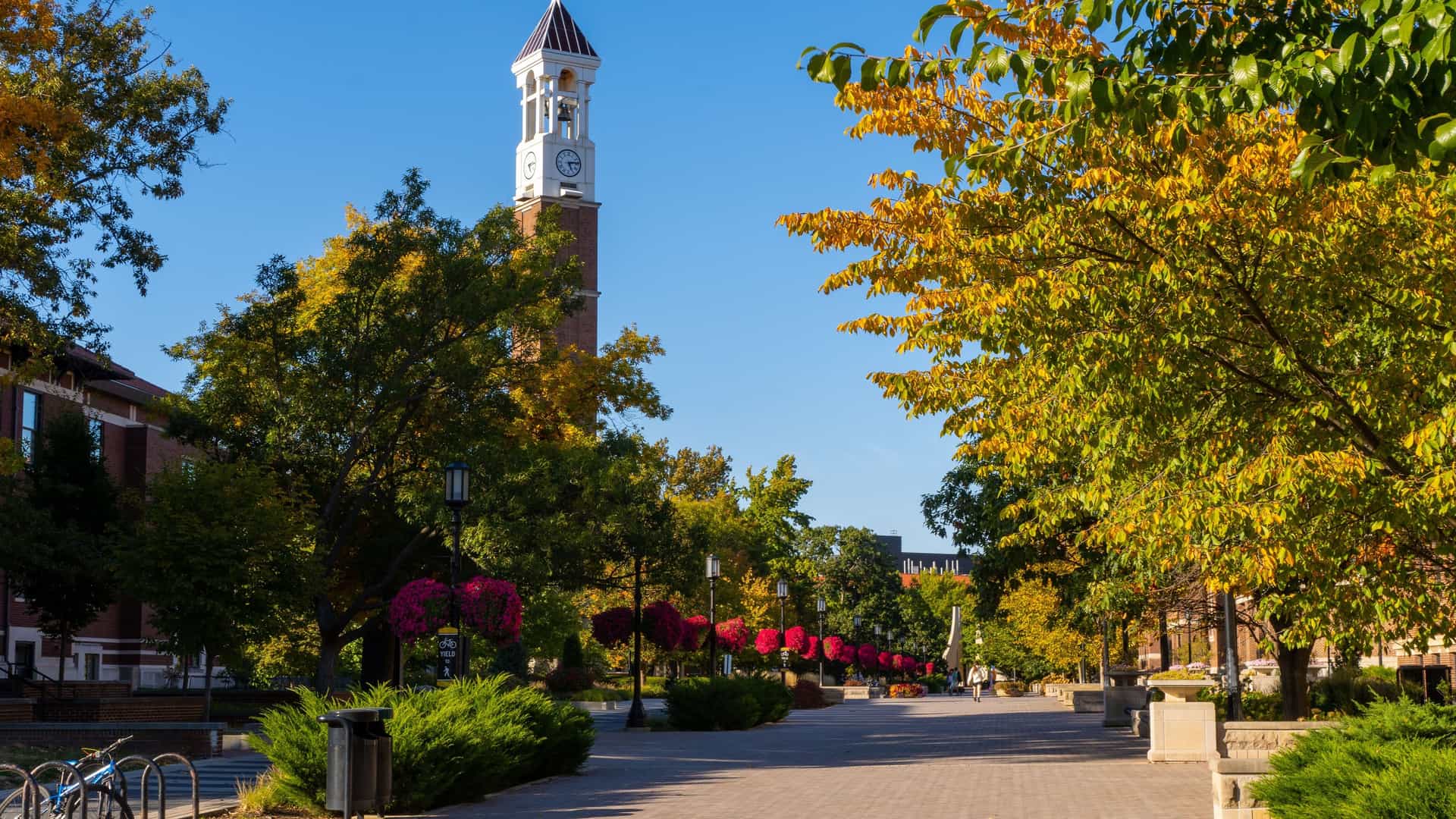"A vibrant autumn scene on the campus of Purdue University in Lafayette, Indiana, showing the iconic Bell Tower and a path lined with trees in full fall foliage. "