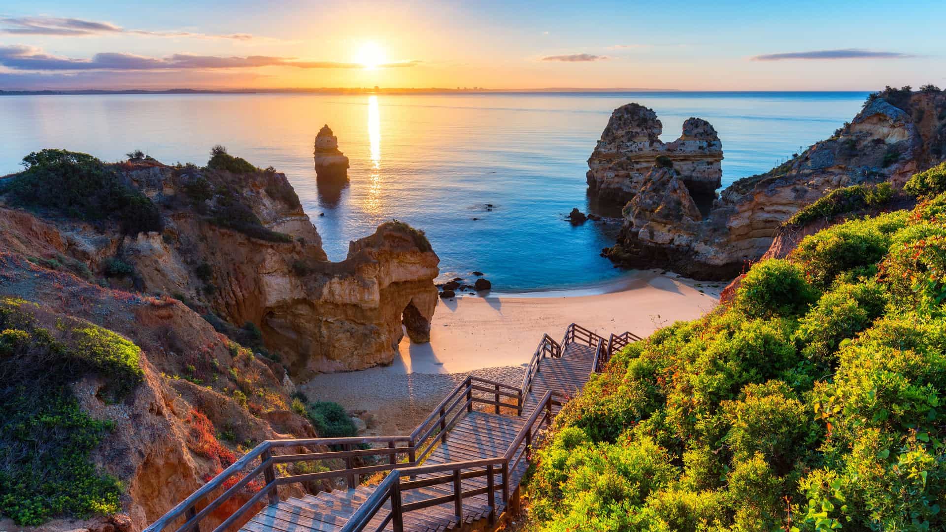 A scenic view of Praia do Camilo beach in Lagos, Portugal, with a wooden staircase leading down to a hidden cove and stunning sea stacks at sunrise.
