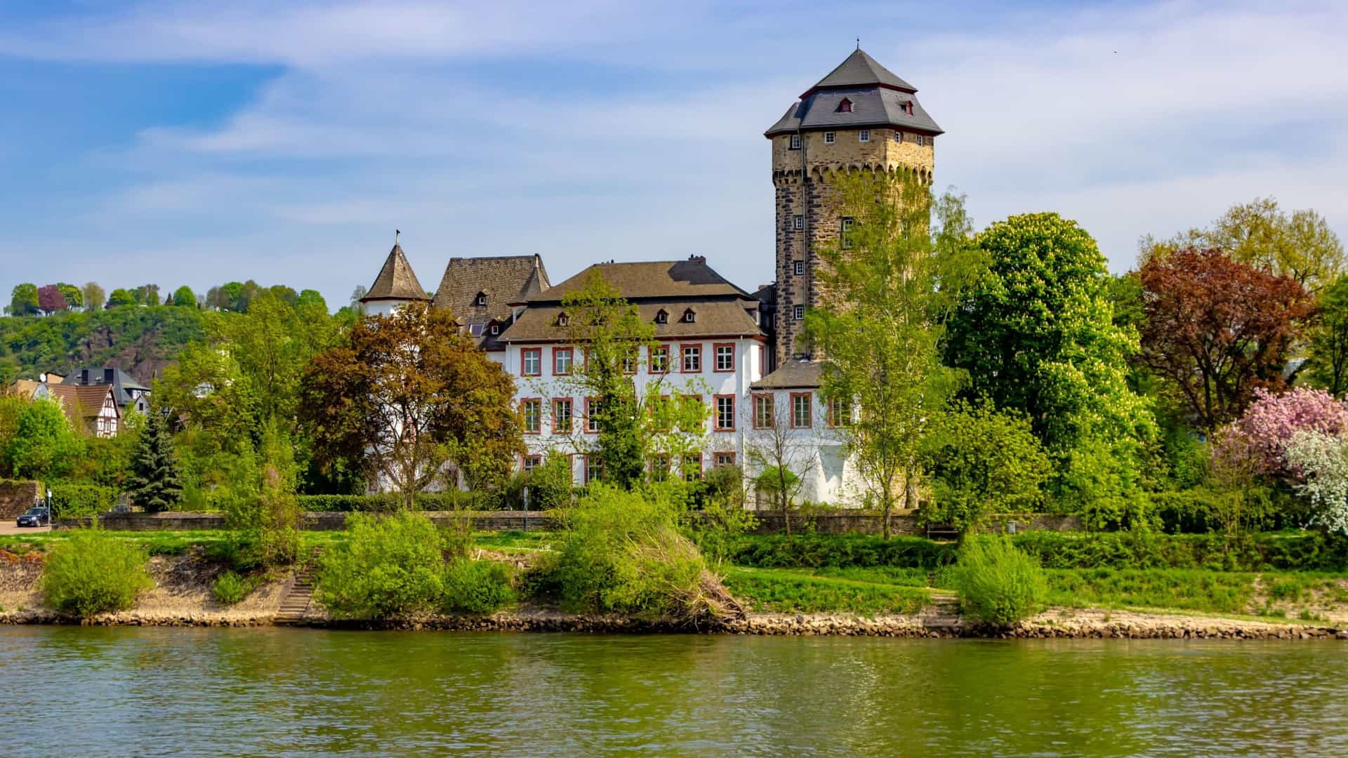 A scenic view of Martinsburg Castle in Lahnstein, Germany, a historic fortress with a stone tower and a half-timbered main building, set amongst green trees by a river.
