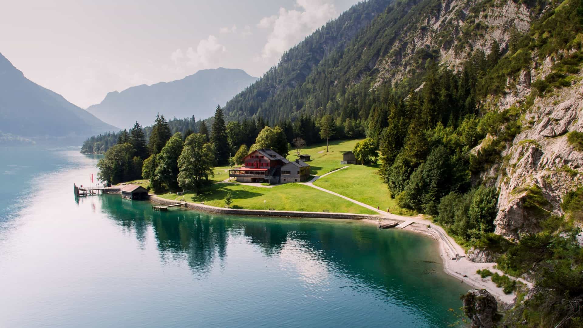 A scenic aerial view of a red-roofed house on the shore of Lake Achen, with a small dock and lush green lawn surrounded by towering mountains and pine forests.