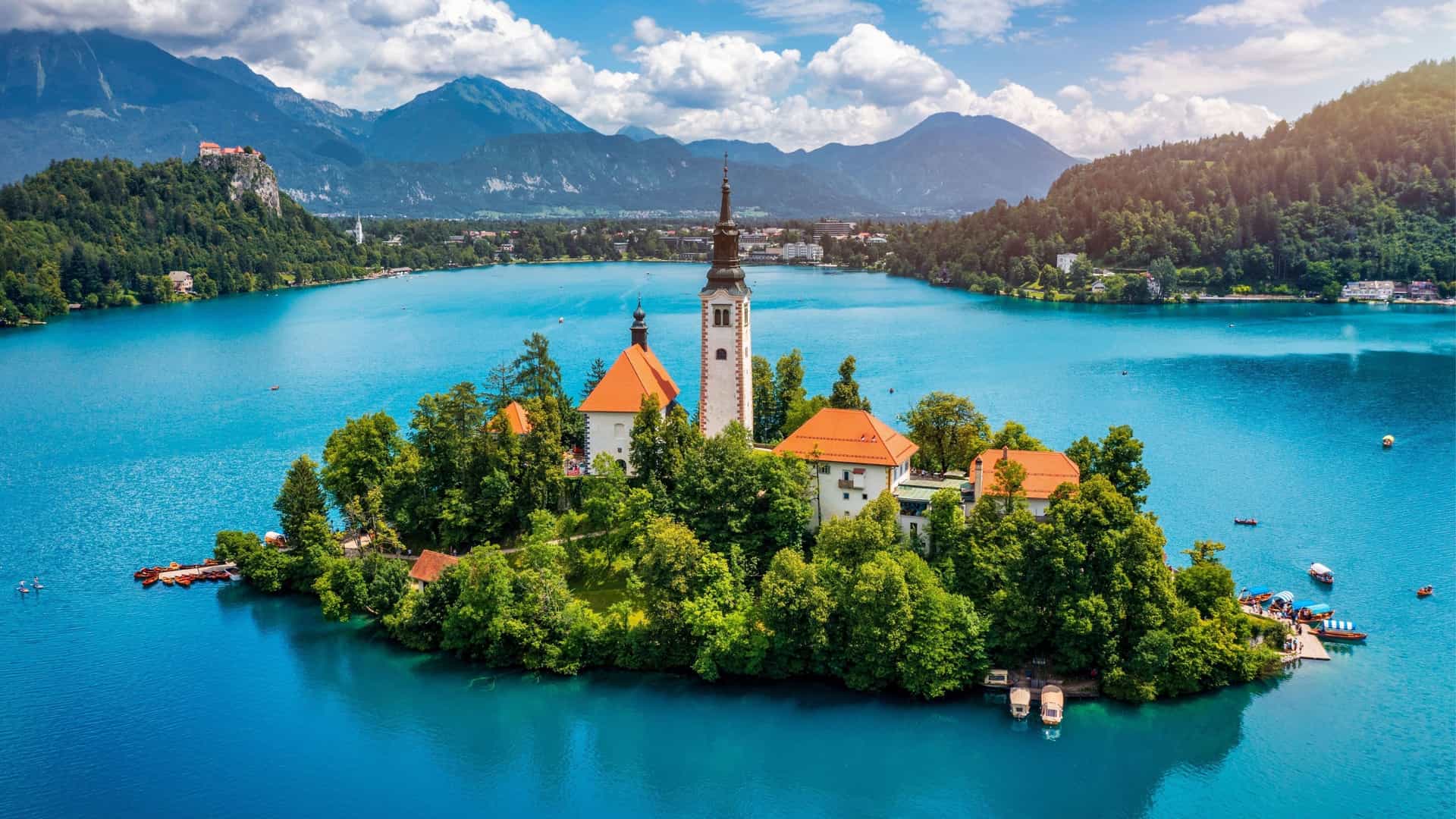 An aerial view of Bled Island and its iconic church steeple, surrounded by the vibrant turquoise water of Lake Bled, with a majestic mountain range in the background.