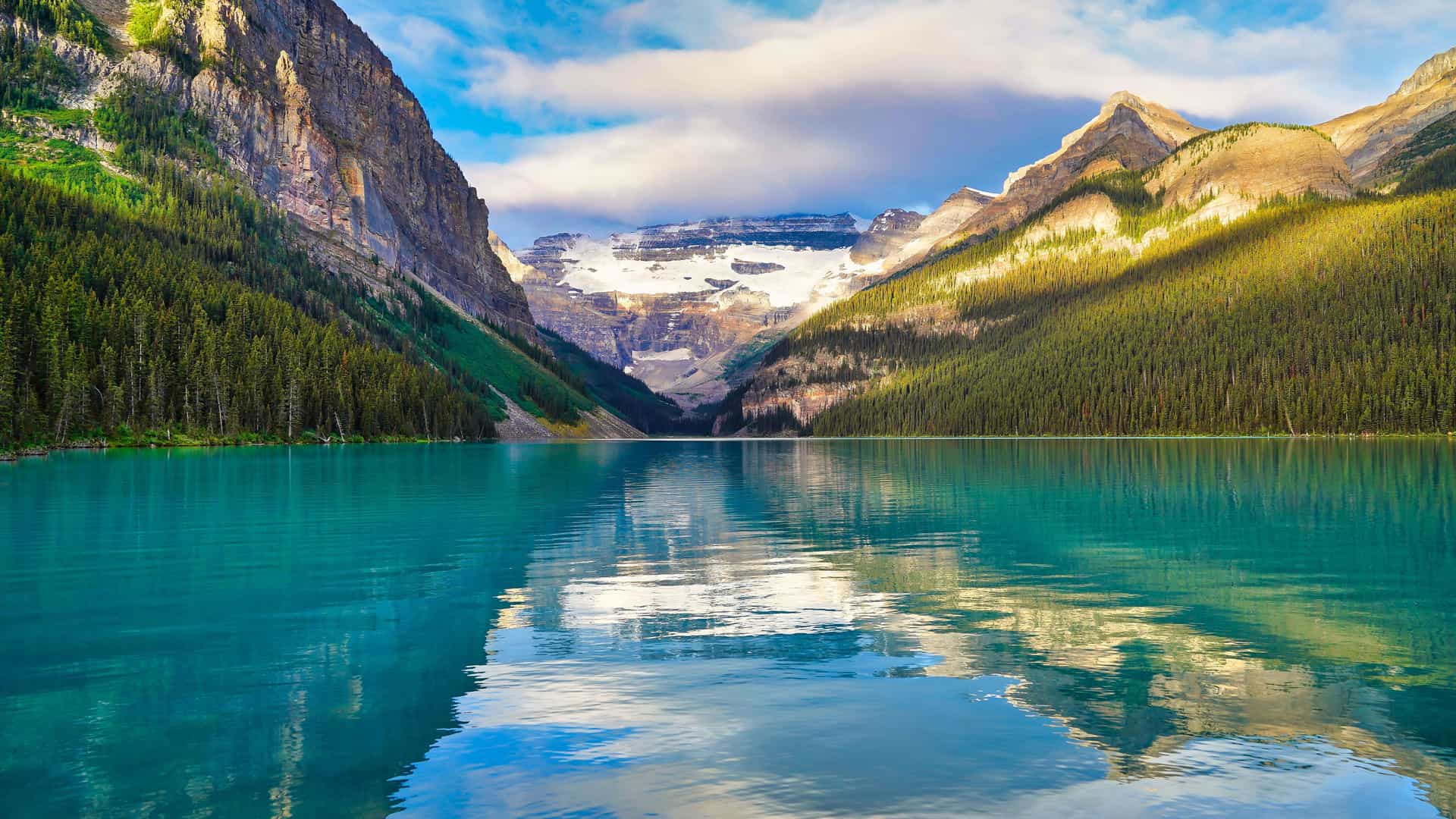 "A picturesque view of the serene turquoise waters of Lake Louise, surrounded by towering, forested mountains and the snow-capped Mount Victoria in the distance in Banff National Park, Canada. "