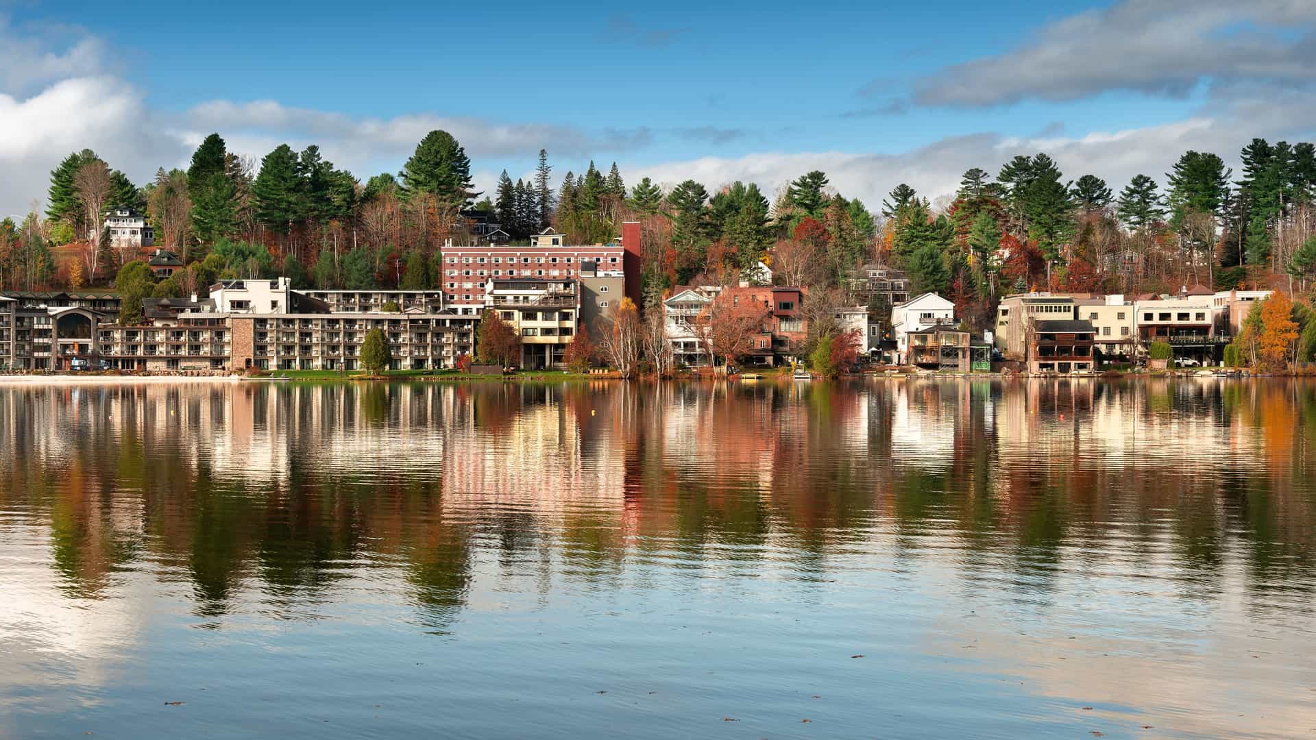 "A serene view of Mirror Lake in Lake Placid, New York, with hotels and houses on the shoreline reflecting in the still water, surrounded by vibrant fall foliage and green evergreen trees. "