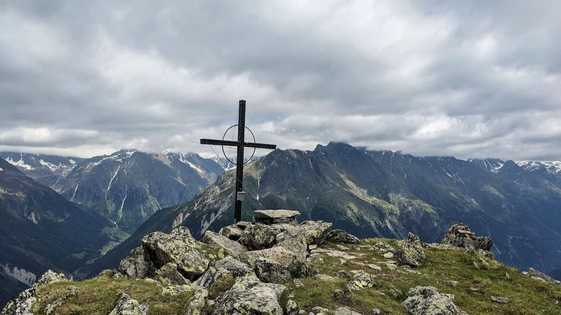 A dramatic view from a rocky mountain summit near Längenfeld, Austria, featuring a large, dark metal summit cross standing in the center. The background shows a vast panorama of steep, green and gray mountains, with snow patches visible on the higher peaks, all under a heavily clouded sky.