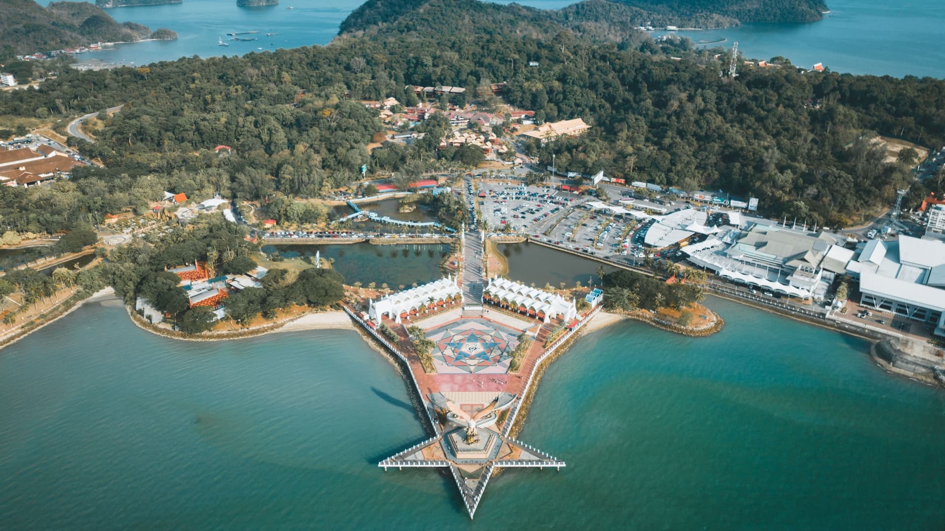 An aerial shot of Dataran Helang, also known as Eagle Square, in Langkawi, Malaysia, with a star-shaped pier extending into the turquoise waters of the harbor, surrounded by forested hills.