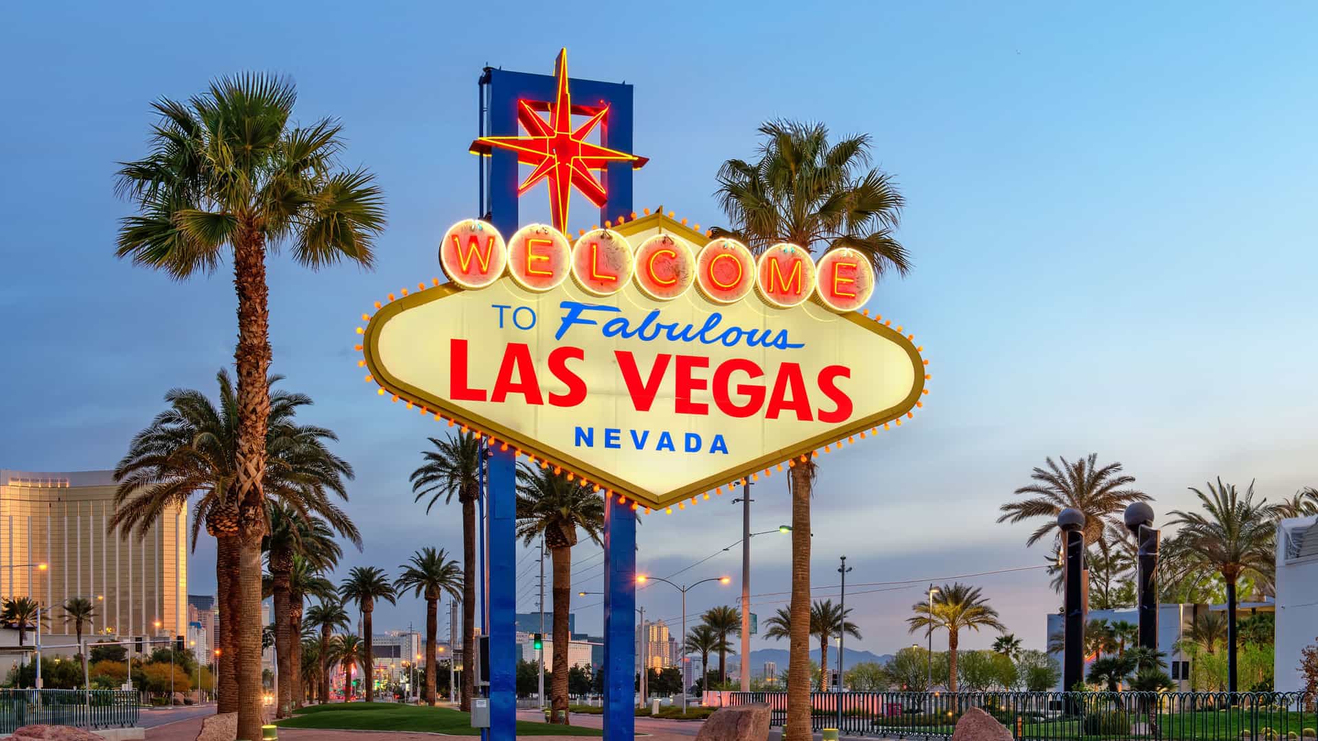 "A wide-angle evening shot of the iconic Welcome to Fabulous Las Vegas sign illuminated with neon lights, surrounded by palm trees against a beautiful blue and pink twilight sky. "