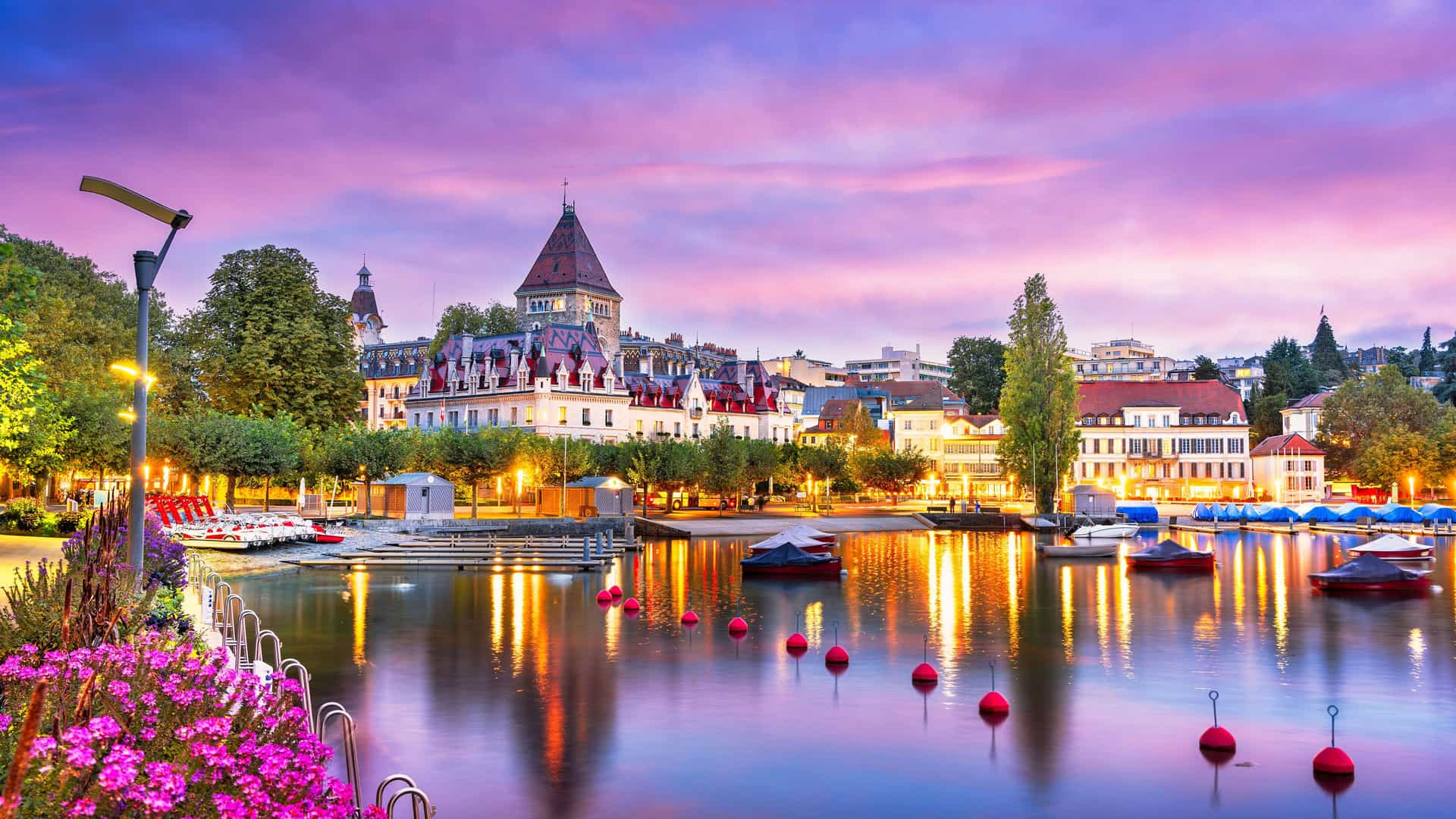 "A breathtaking sunset shot of the waterfront in Lausanne, Switzerland, with the city's charming architecture reflecting in the still waters of Lake Geneva, framed by pink flowers and moored boats. "
