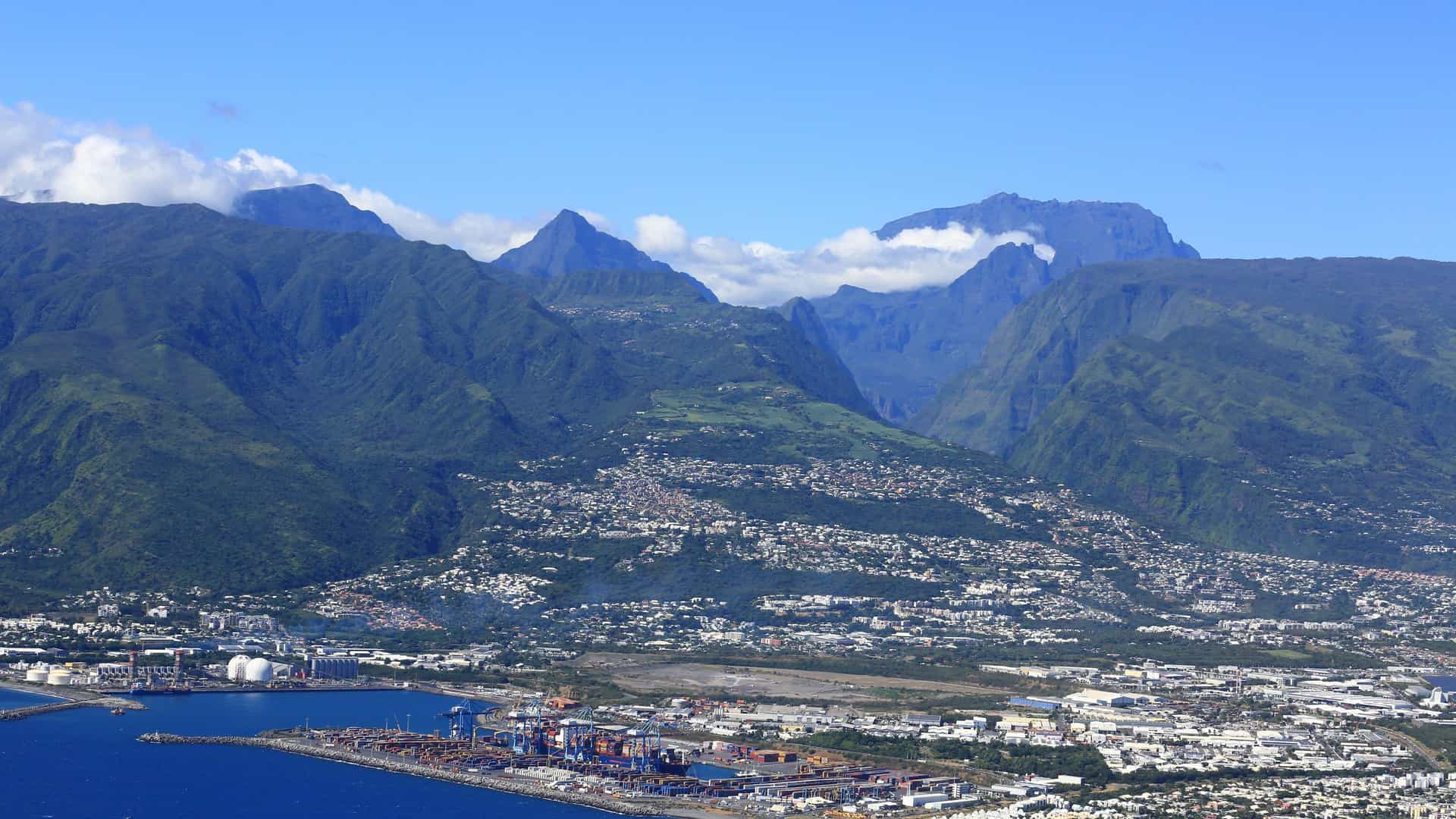 An aerial view of Le Port, a bustling harbor city in Réunion, with a busy industrial port in the foreground, and the dramatic, lush green mountains of the island's interior rising in the background under a blue sky.