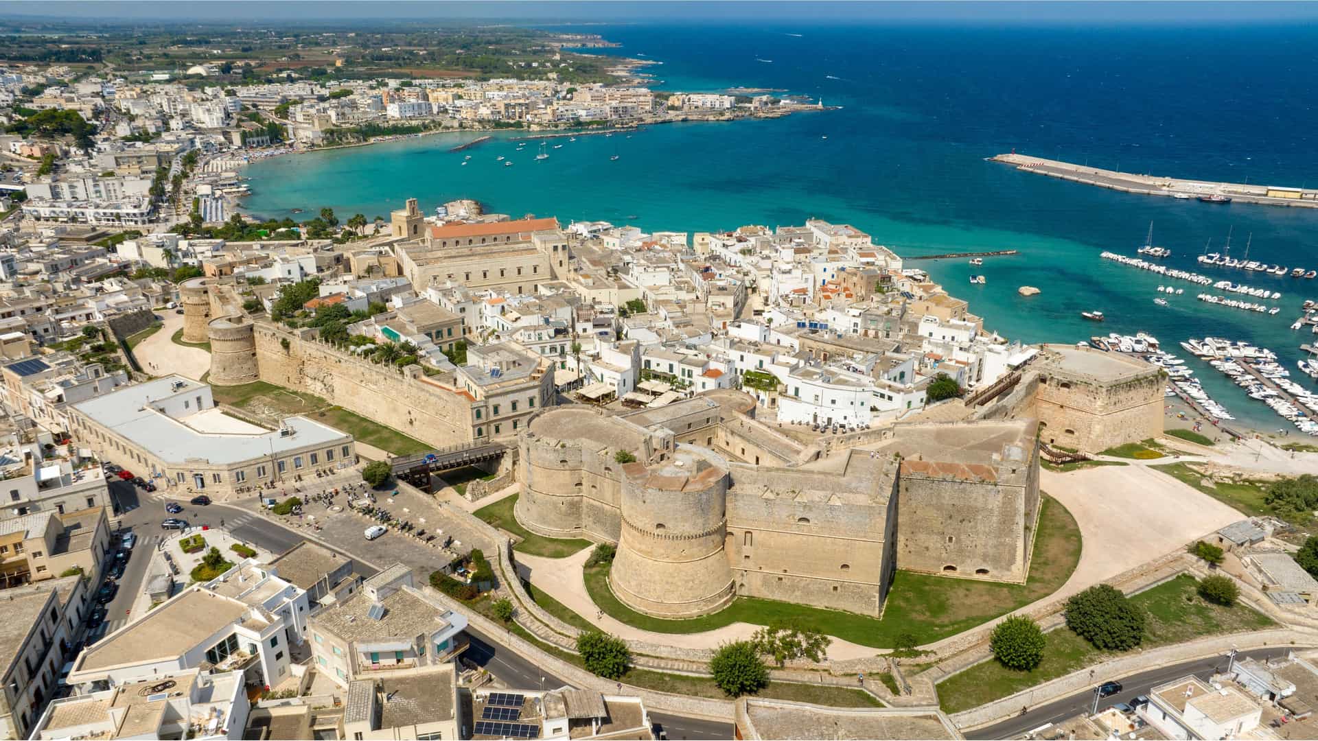 "An aerial view of the historic town of Otranto in the province of Lecce, Italy, showcasing the large coastal castle and the marina with boats in the turquoise sea. "