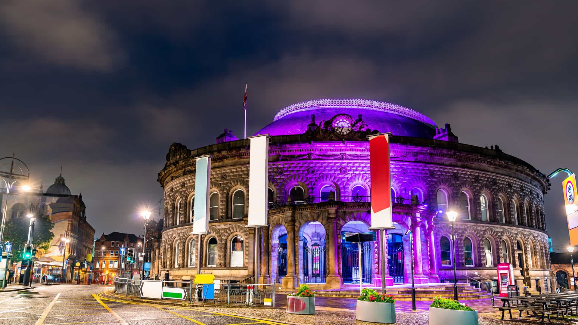 "A long exposure night shot of the Leeds Corn Exchange in Leeds, England, illuminated with vibrant purple lights under a cloudy sky. "