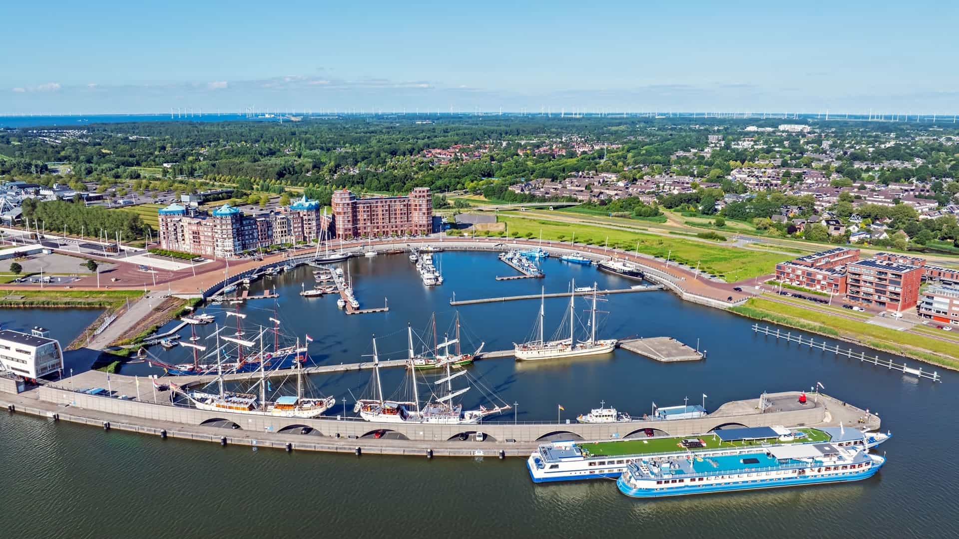 An aerial view of a semi-circular harbor filled with large sailing ships and modern boats, bordered by brick apartments and green forests.