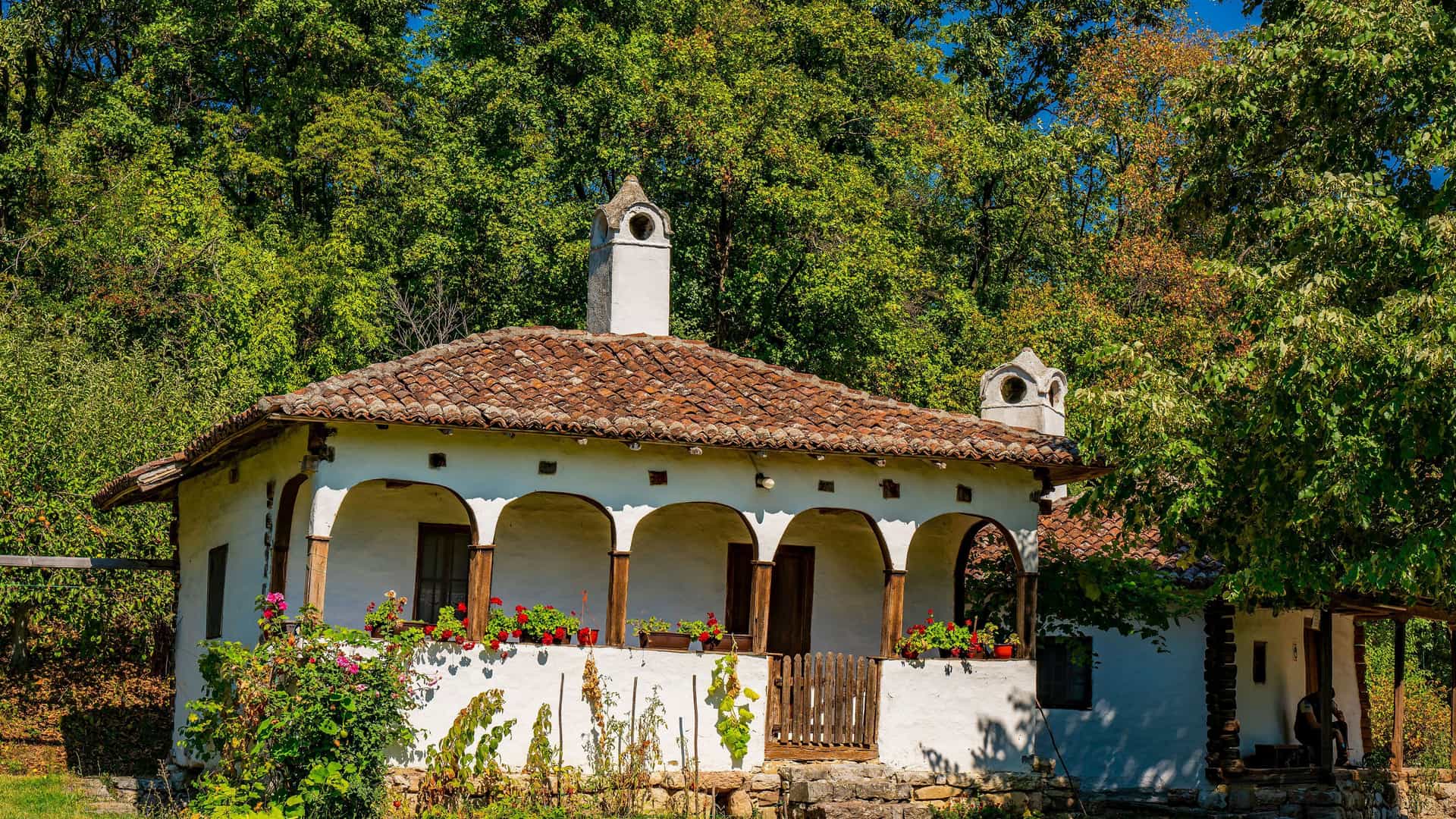 A rustic, white, traditional Serbian house with a red tile roof and a covered porch supported by wooden pillars, surrounded by dense green trees.